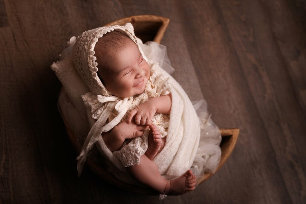 A newborn baby giggles while curled up in a basket while wearing a lace dress and bonnet 3d ultrasounds Toronto