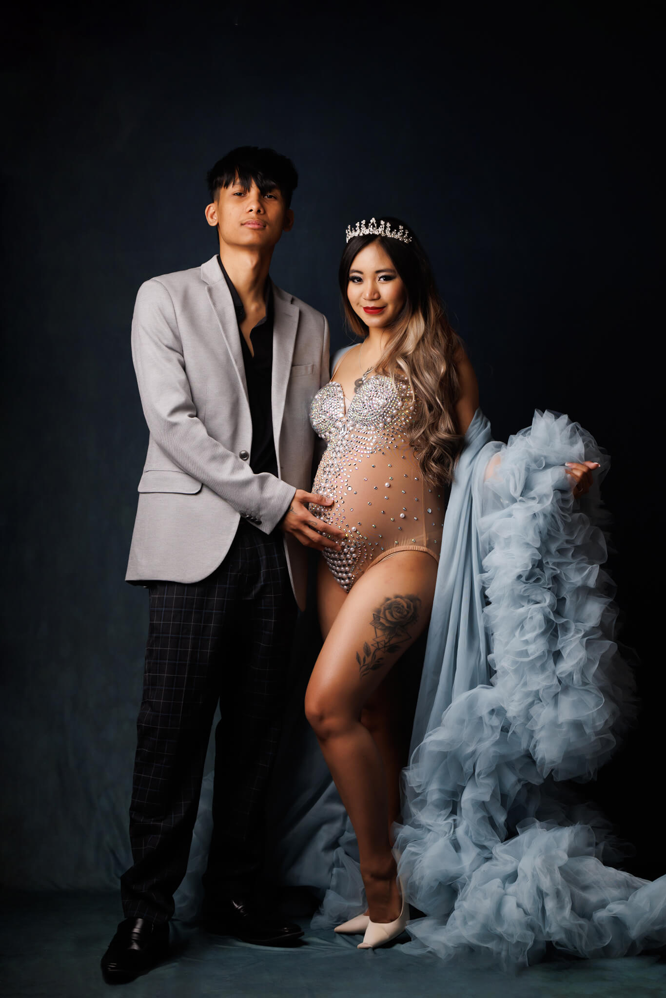 A mother to bee stands in a studio in a tule gown and bedazzled maternity one-piece while her partner stands with a hand on the bump Oona Cares