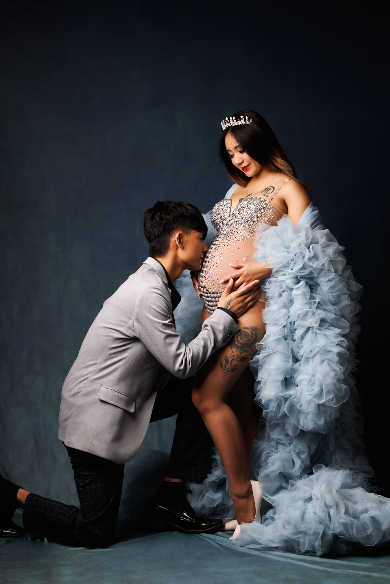 A mother-to-be in an ornate luxury maternity gown and crown stands in a studio while the father-to-be kneels and kisses her belly Oona Cares