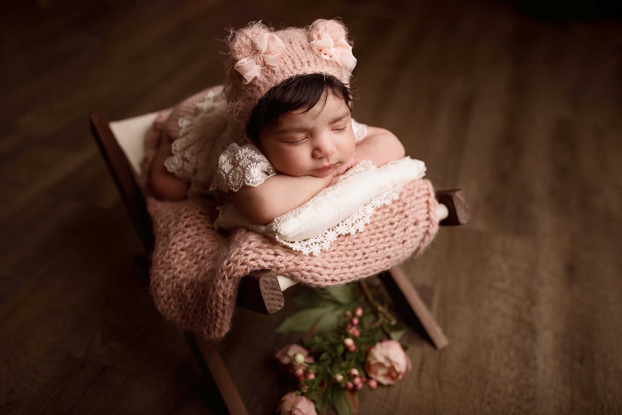 Newborn baby wearing a white lace dress and pink knit hat sleeps on a wooden chair Toronto Baby Show