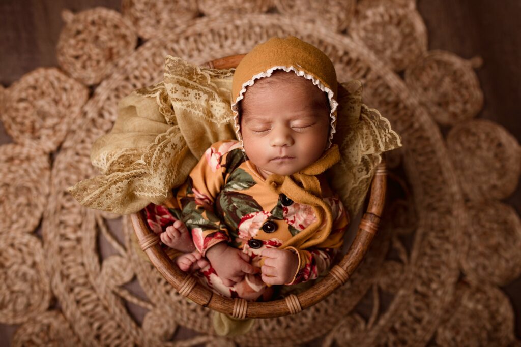 newborn sleeps in a basket on a woven mat wearing a brown bonnet