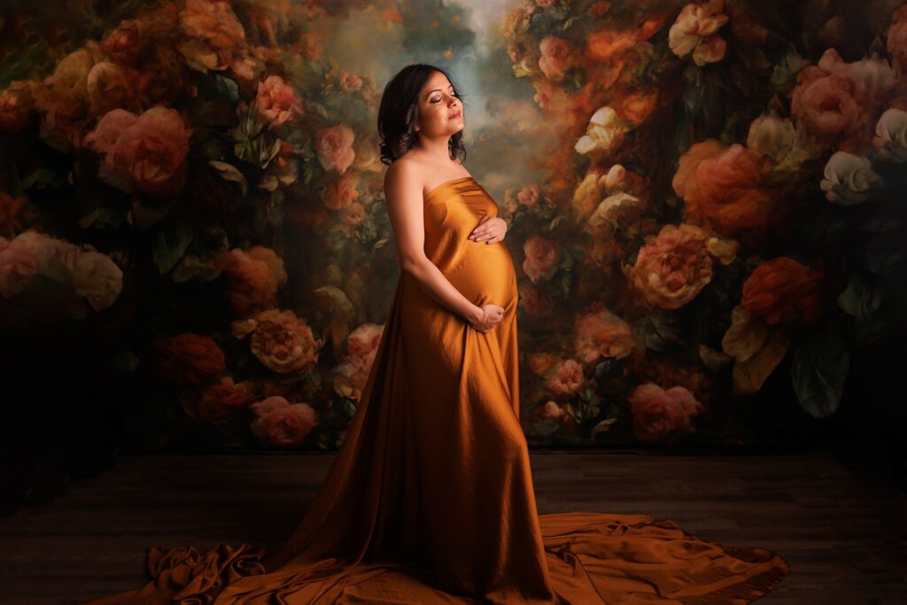 Mother to be wearing a long gold maternity gown stands in a studio in front of a carnation backdrop Toronto Birthing Centre
