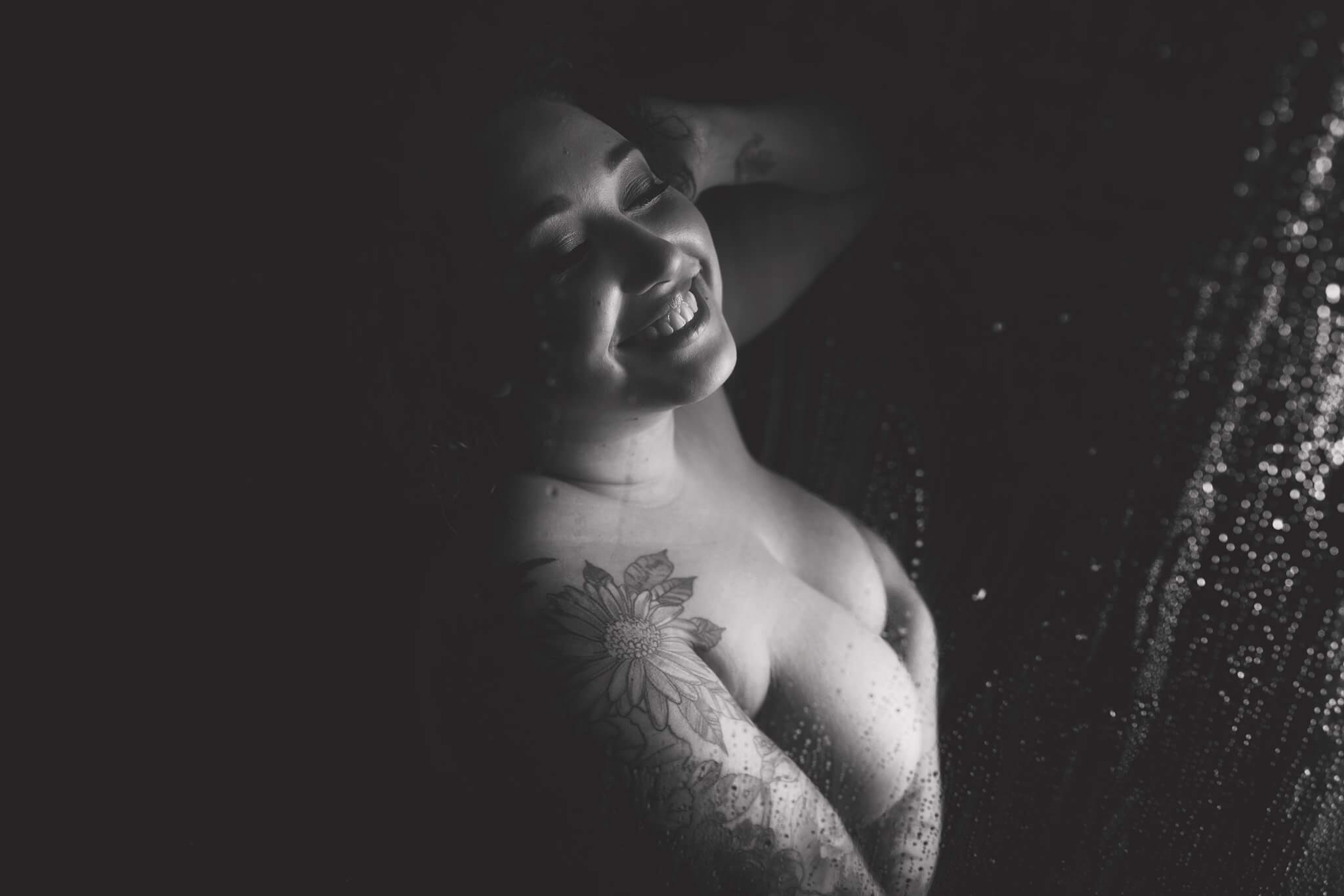 Photography by Toronto Photographer Shanna Parker Black & White photo of a woman smiling in a shower with tattoos on her right arm and shoulder