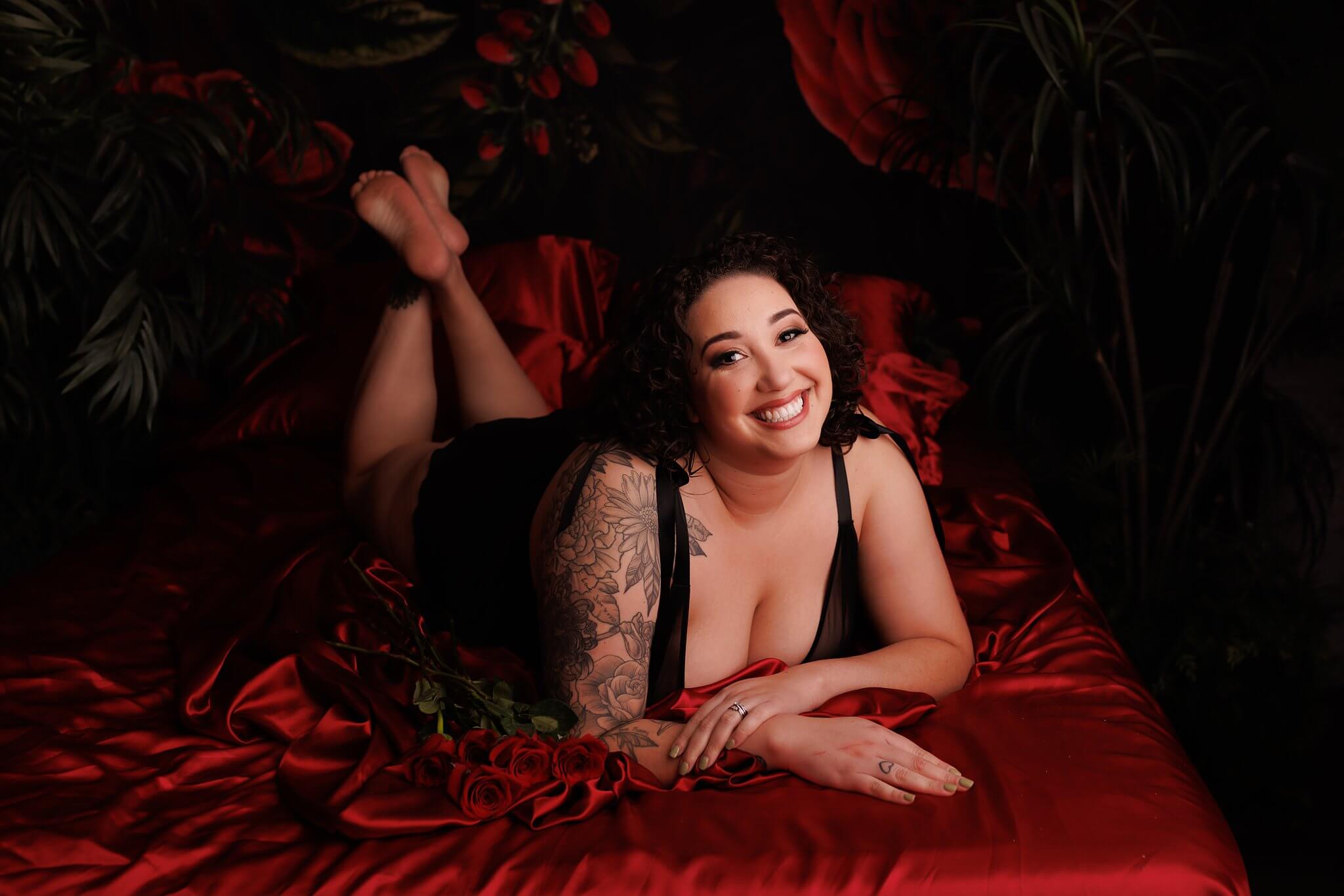 Beautiful woman with tattoos on her right arm, laying in lingerie on red satin sheets, smiling at the camera