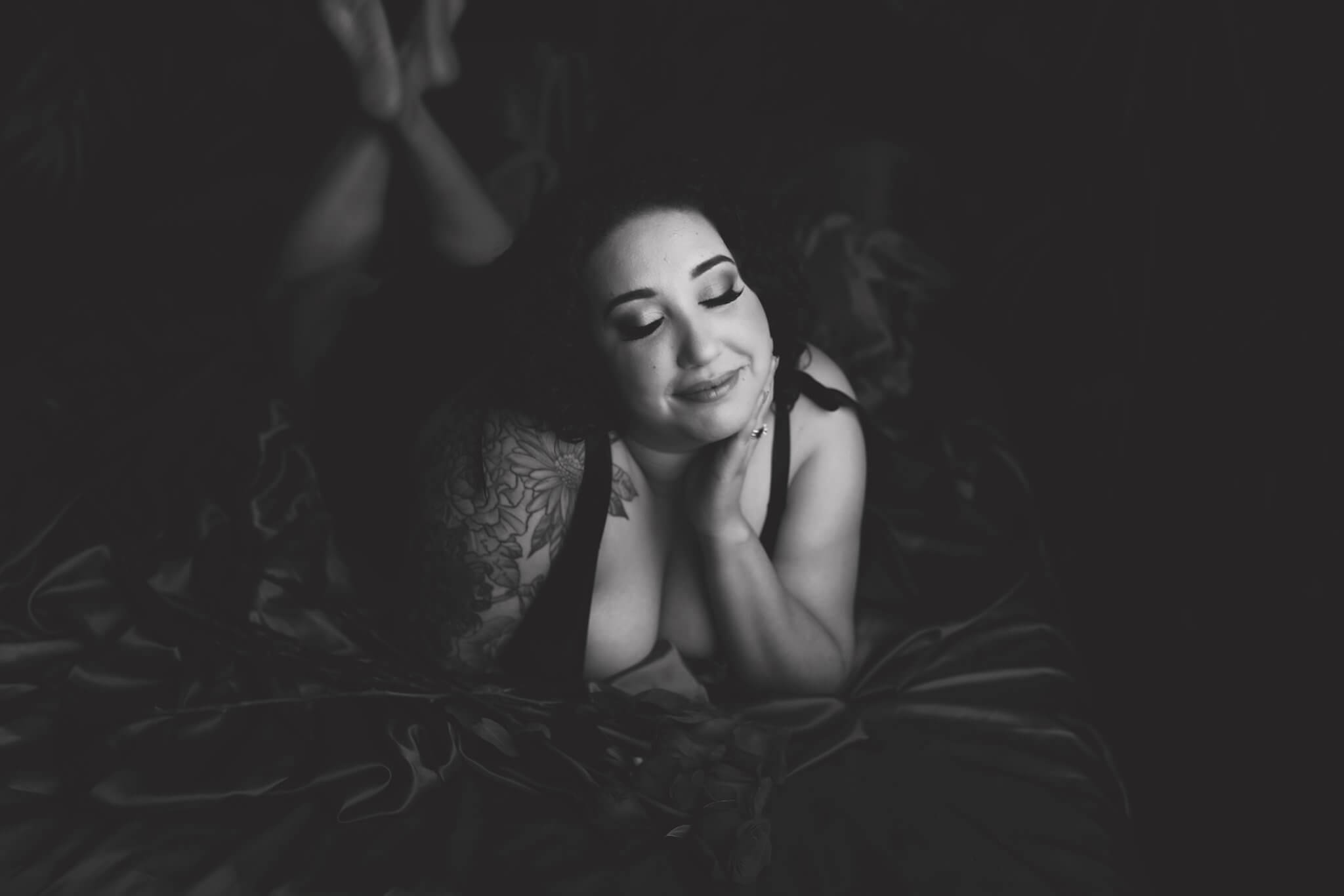 Black & White photo of a woman lying on her stomach in black lingerie on red satin sheets, she has tattoos on her right arm and left leg