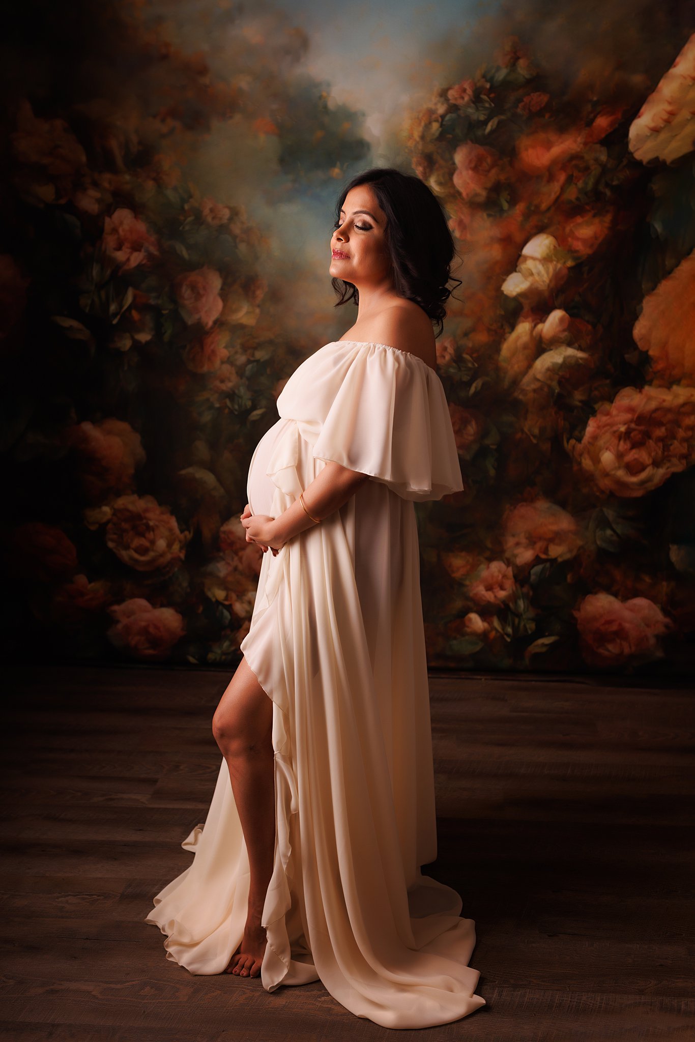 A mom to be holds her hands under her bump standing in a studio with a floral backdrop