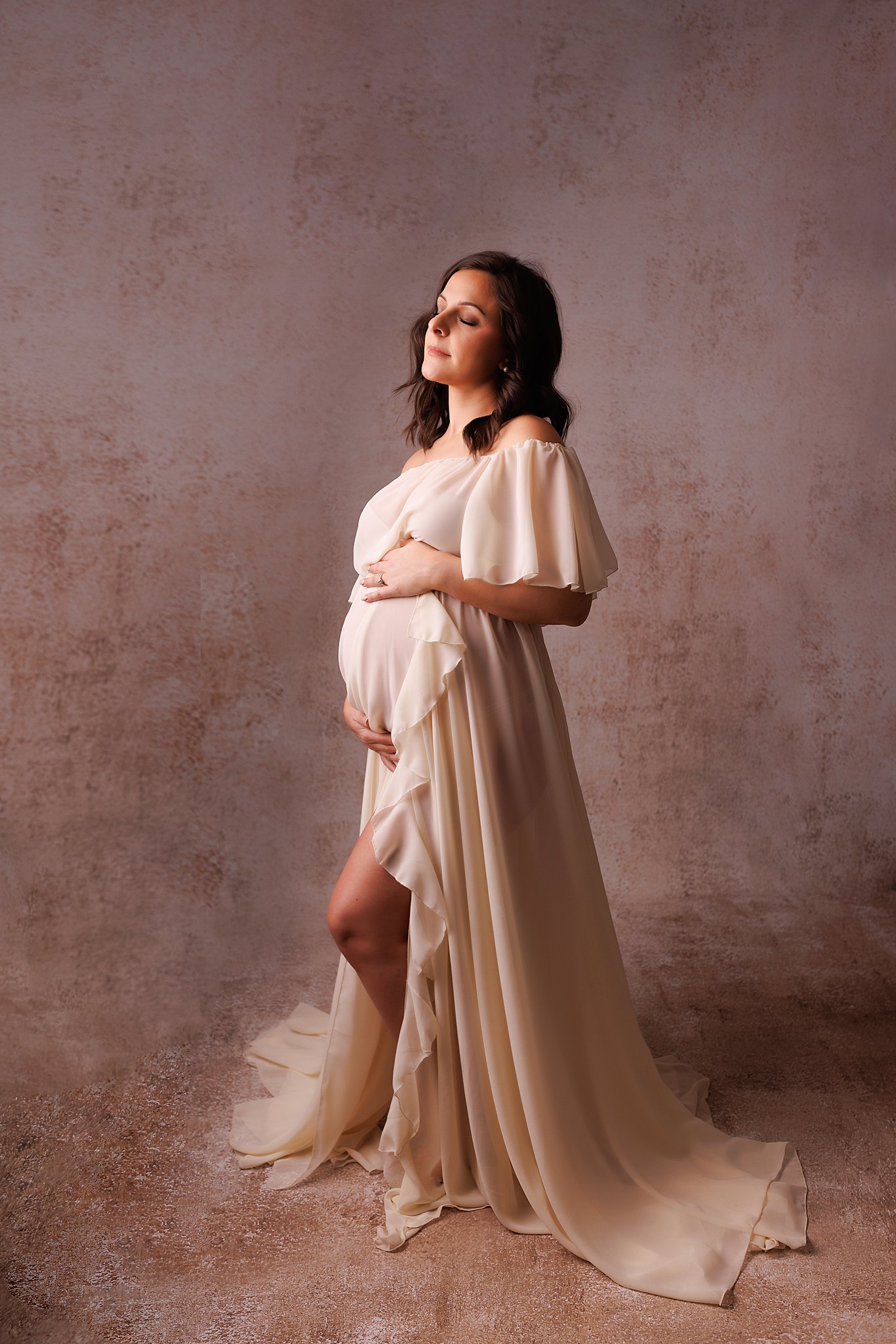 A mother to be stands in a studio holding her bump wearing a long beige maternity gown Toronto Doula