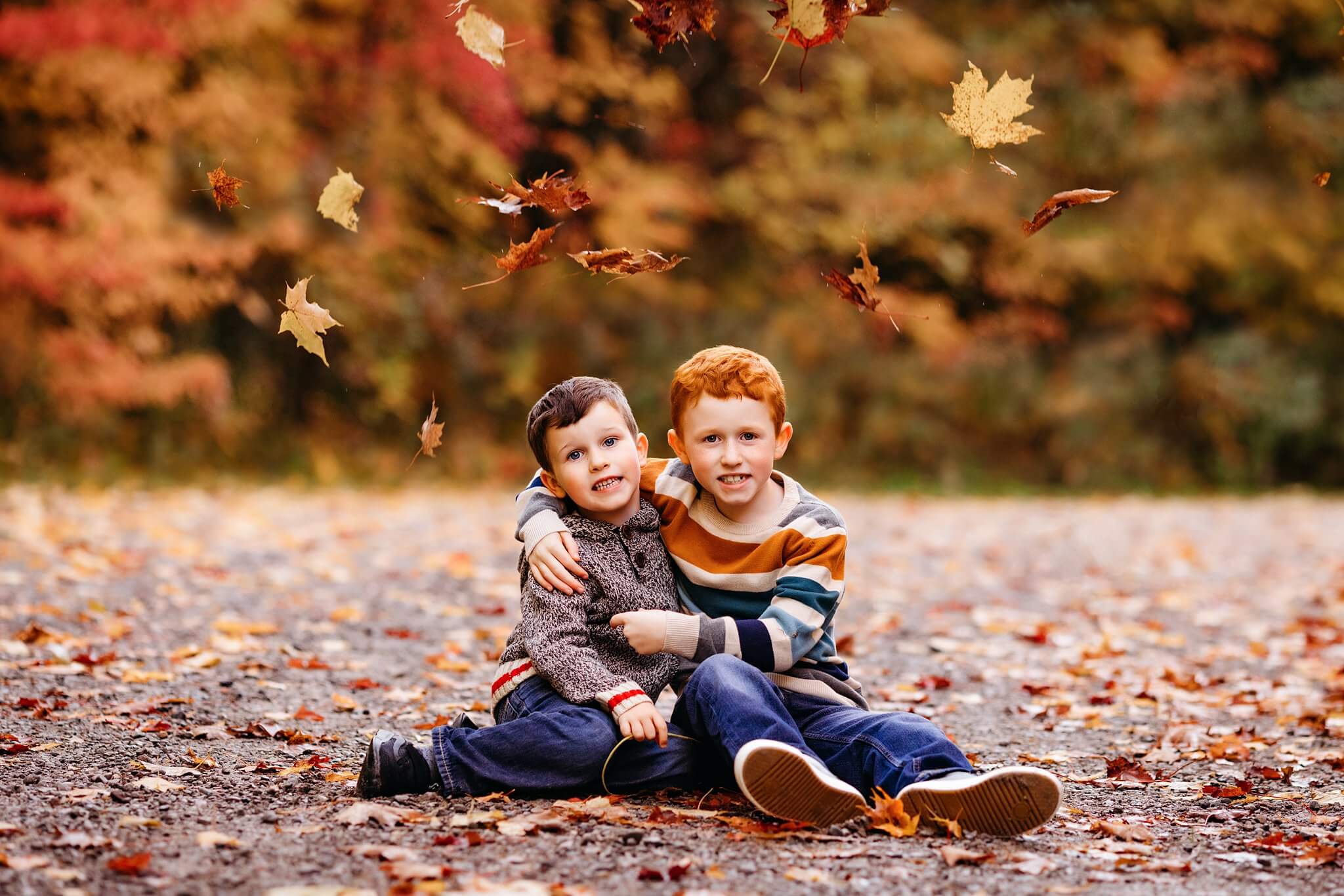 Two small boys sit amongst leaves with leaves falling around them