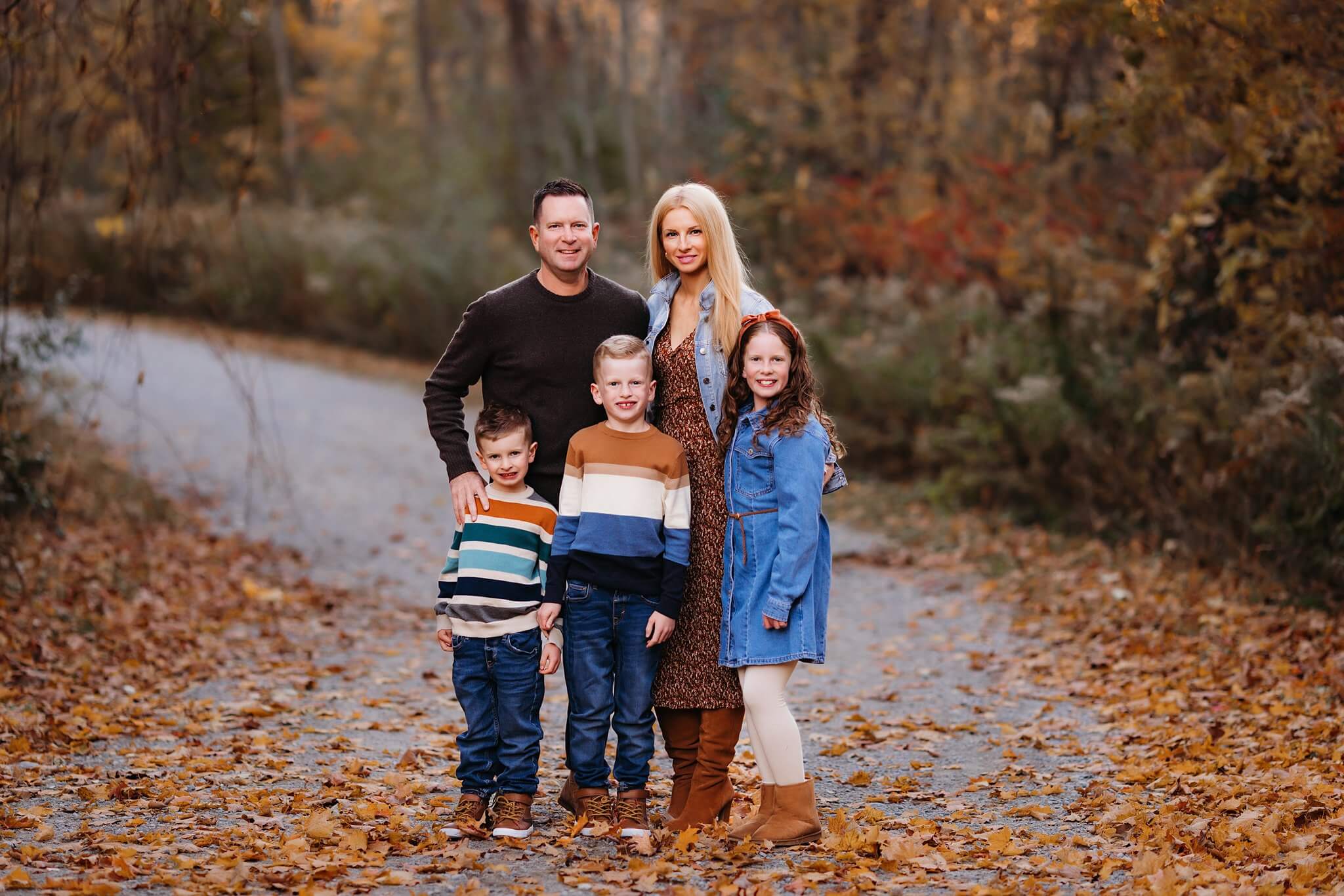 Family with two young kids stand on a leaf covered path amongst fall colors posing for a family photo by Toronto Family Photographer