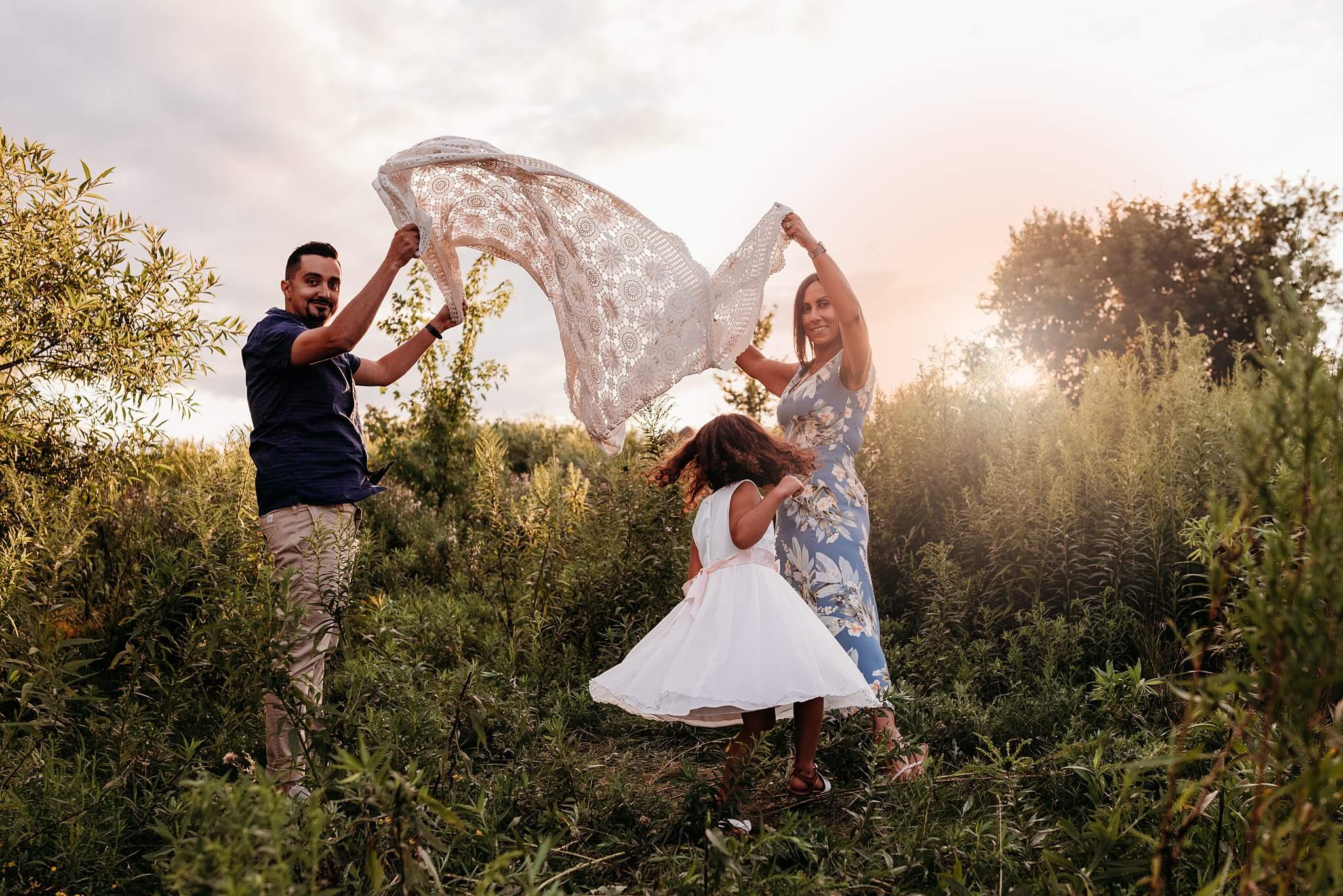 Little girl running under lace blanket her parents are tossing at sunset