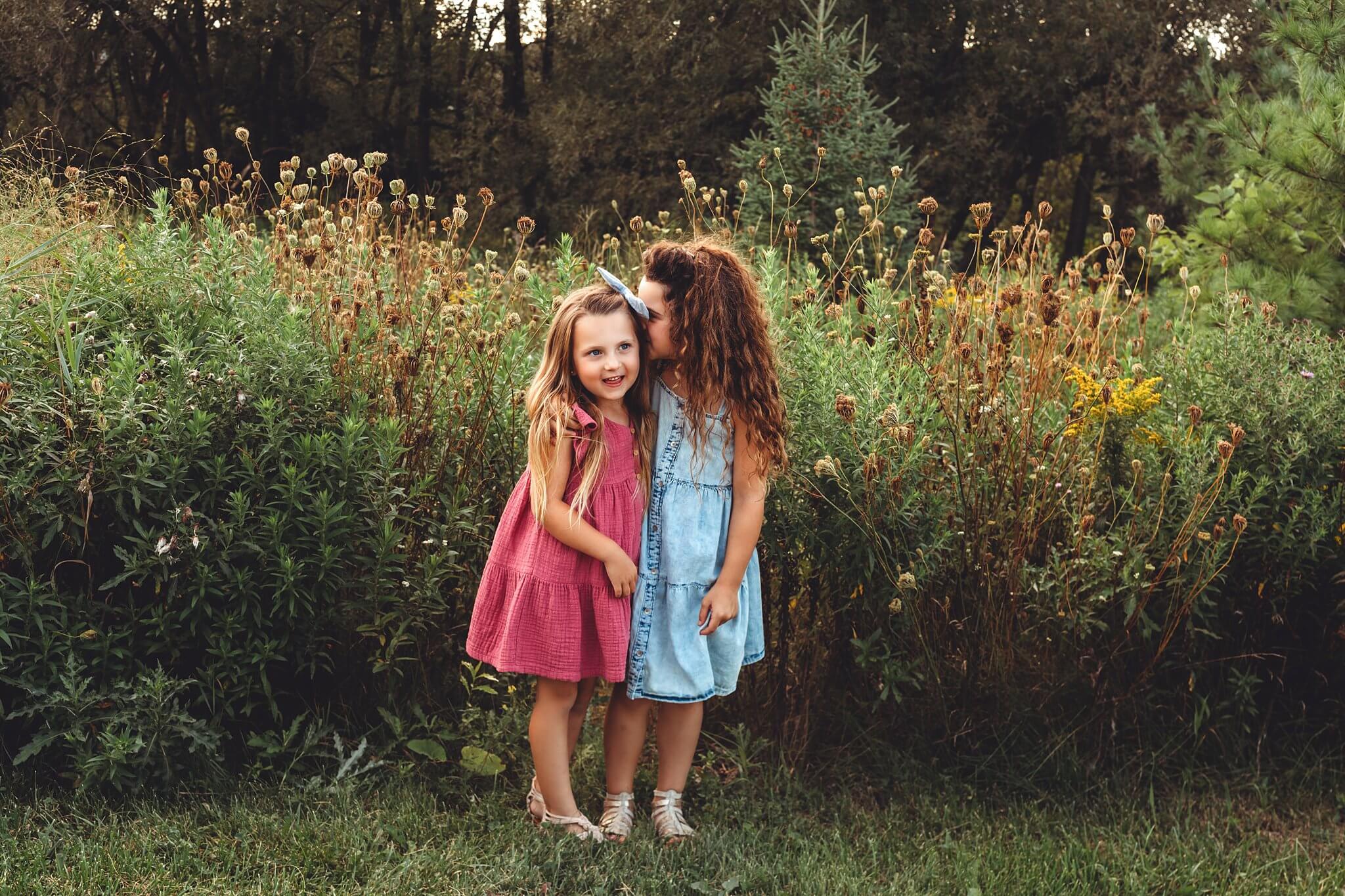 Young sisters in surrounded by tall grasses, both in cute dresses and one is whispering a secret to the other by Toronto Family Photographer