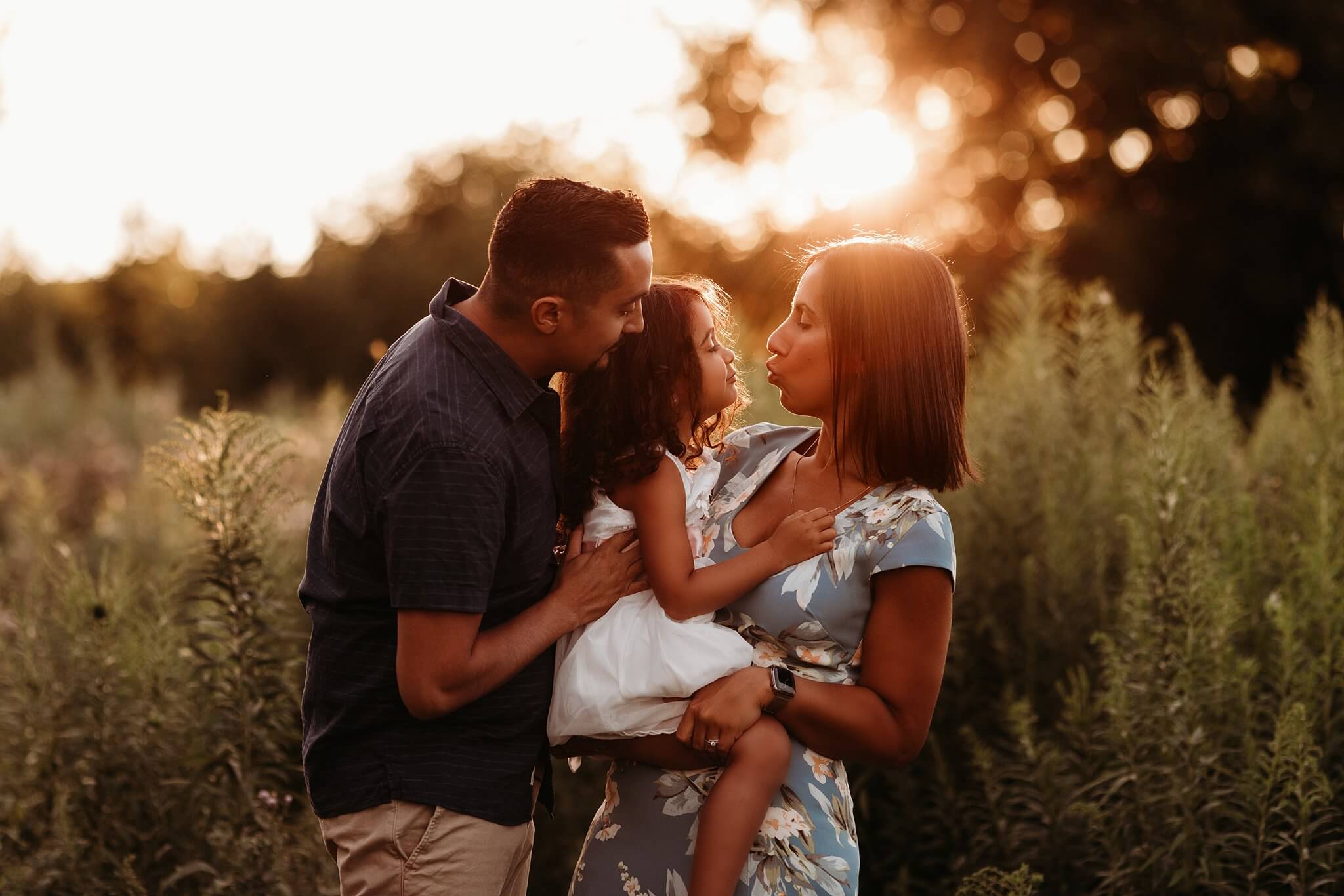 Photo of a Mother and Father holding and snuggling with their daughter at sunset by Toronto Family Photographer