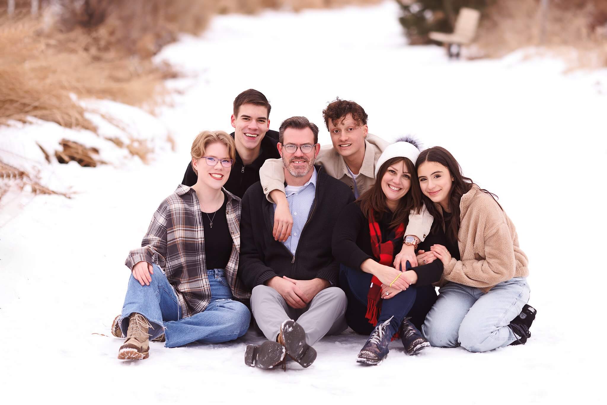 Snowy family photo, parents and four teenage kids sit on a winter path covered in snow