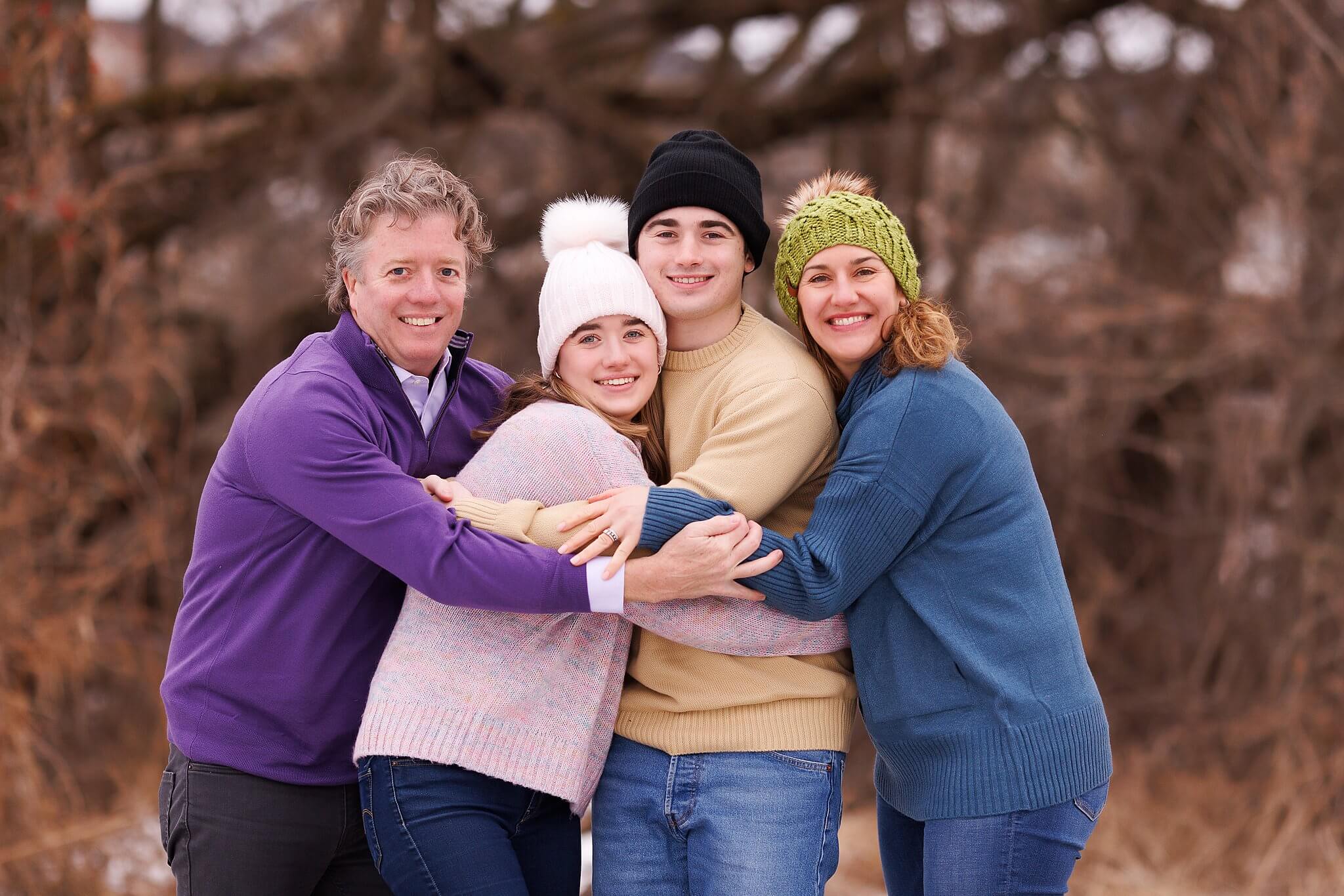 Family in colorful winter hats and sweaters embracing with bare trees in the background. Winter portrait by Toronto Family Photographer