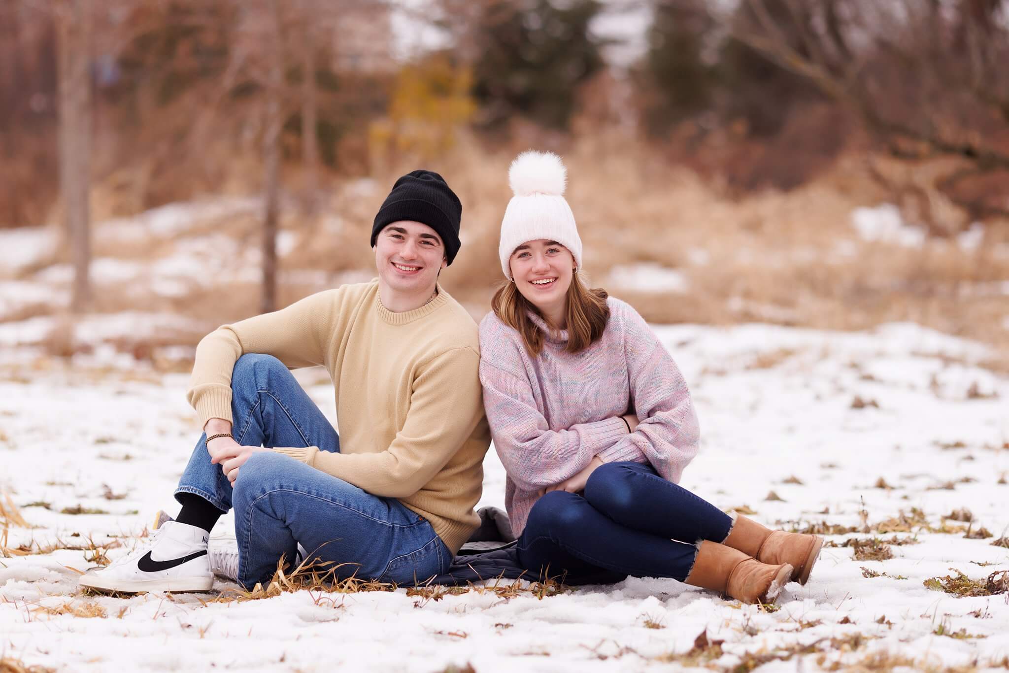 Teenage brother and sister in winter hats and sweaters sit in the snow and smile at the camera