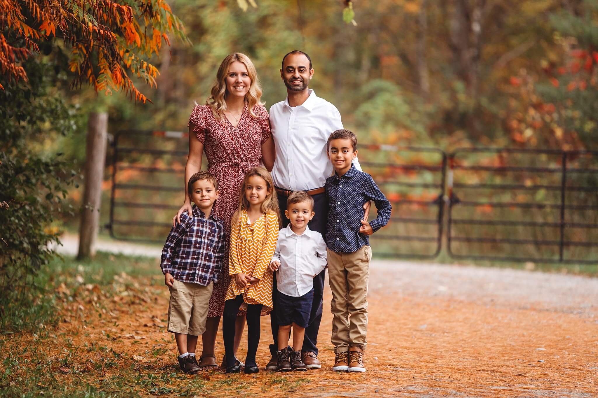 Fall Family Portrait of two parents with their four children standing in front of a fence surrounded in fall leaves