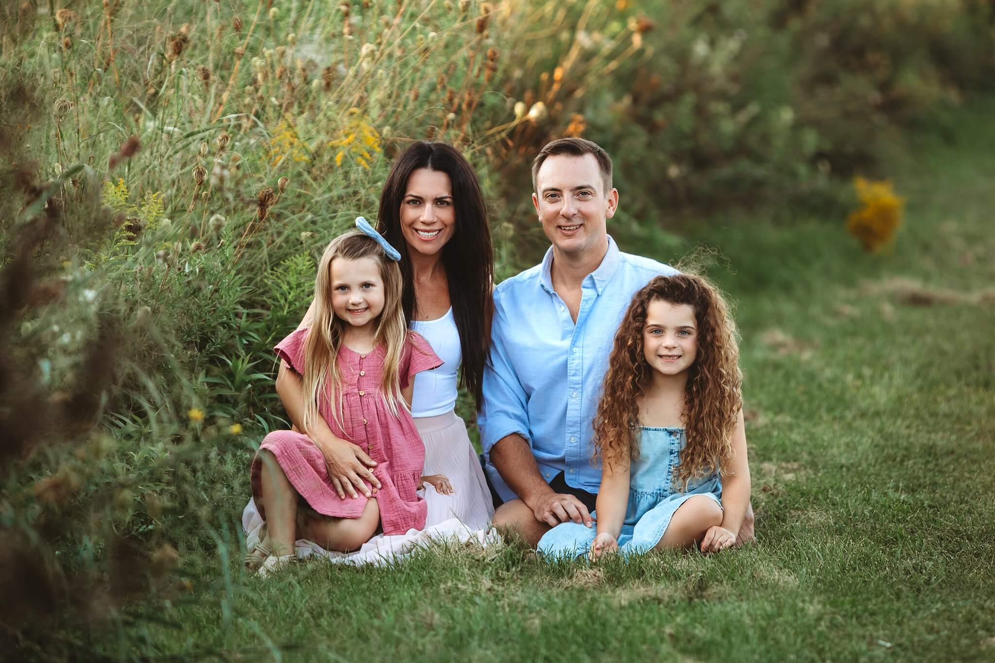 Family with two cute daughters sits on the grass in pastel clothing smiling at the camera by Toronto Family Photographer Shanna Parker