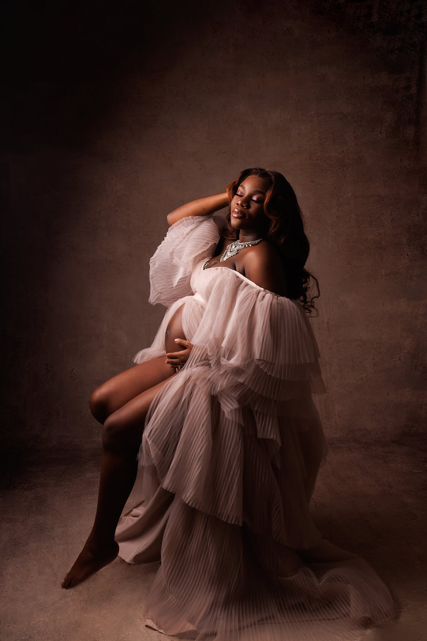 Gorgeous pregnant woman sitting in a flowing taupe gown, hand in her hair