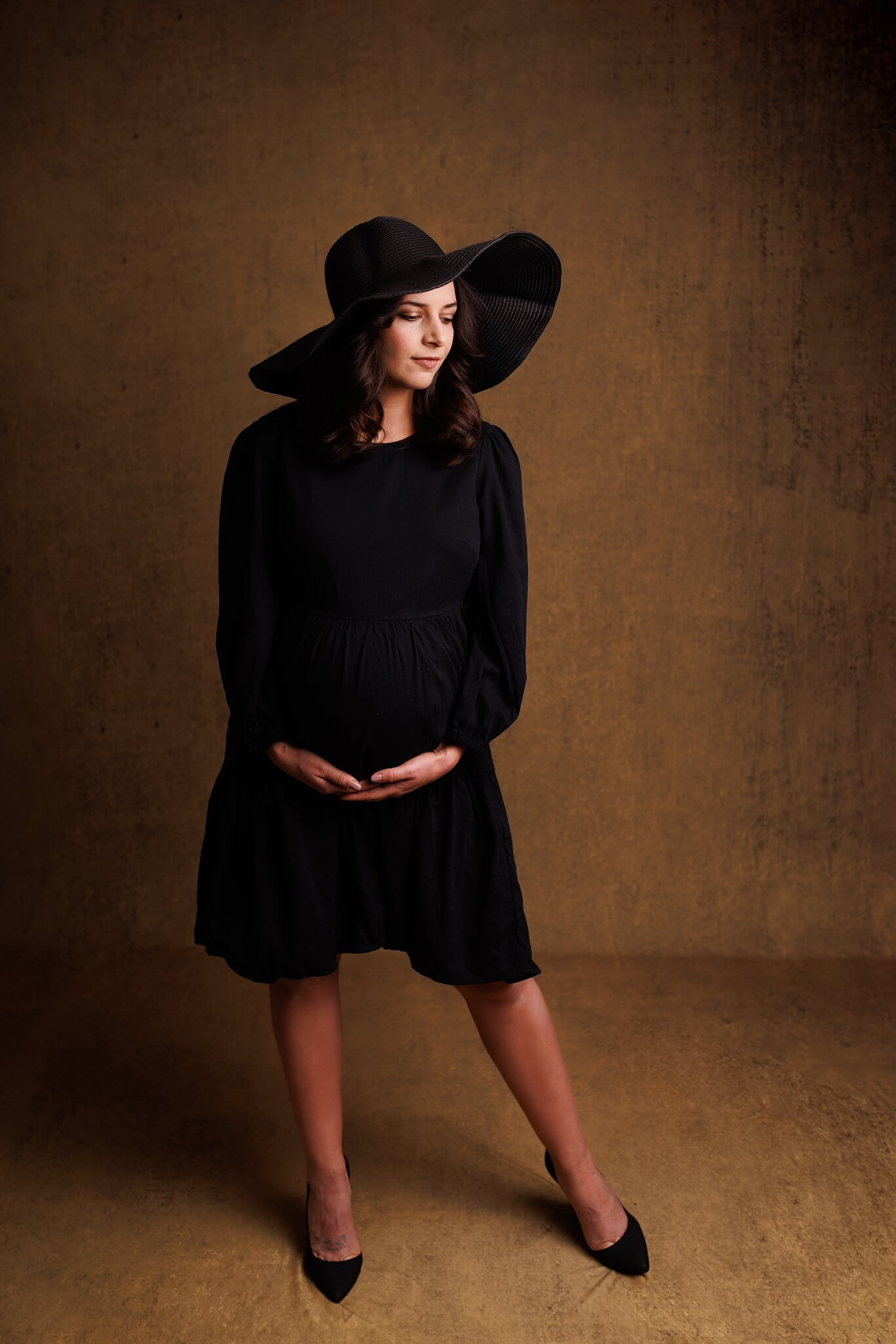 Trendy mama in a short black dress and large black hat holding her pregnant belly by Toronto Photographer Shanna Parker