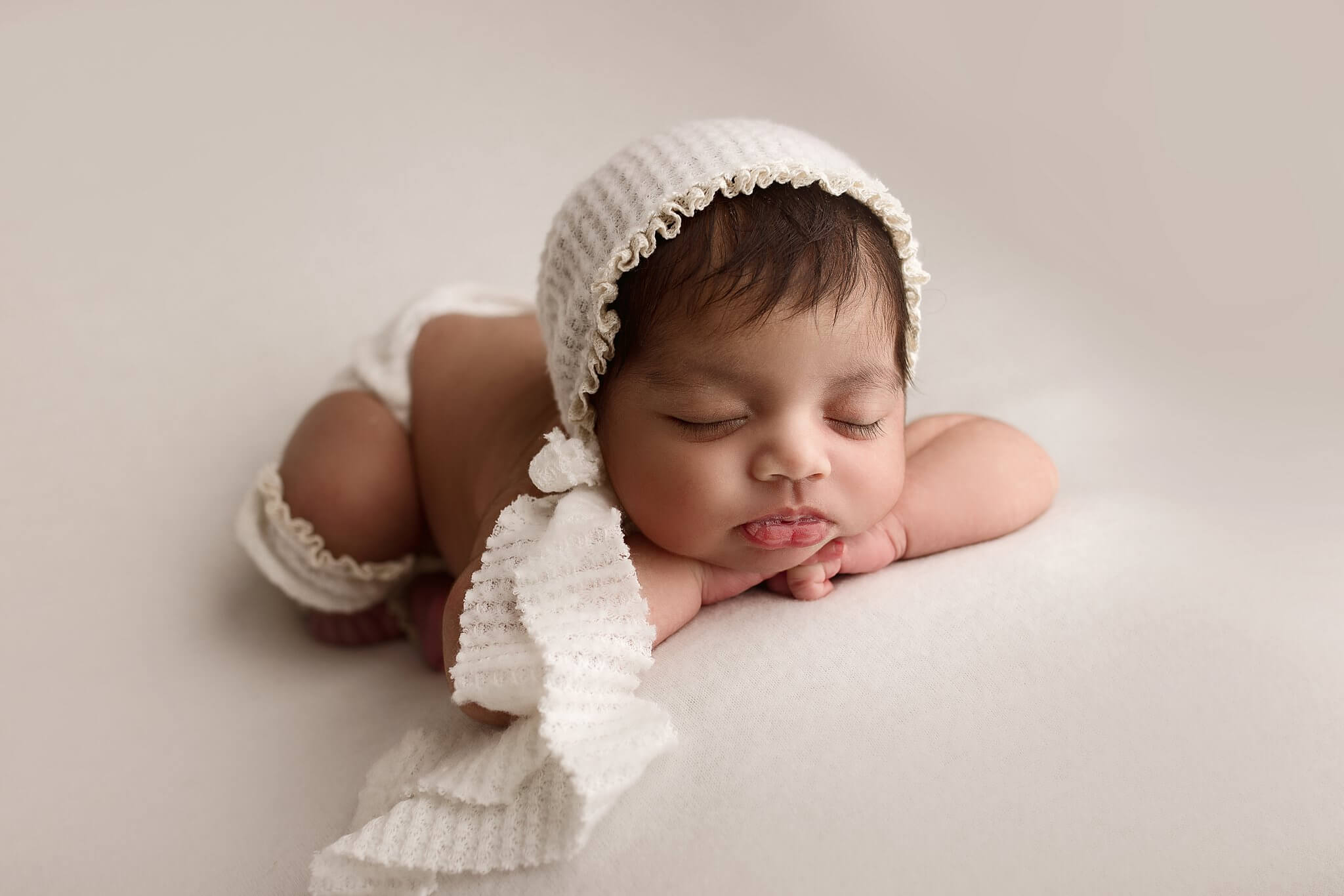 Newborn Baby Girl in a white bonnet and leg warmers laying on a simple white backdrop