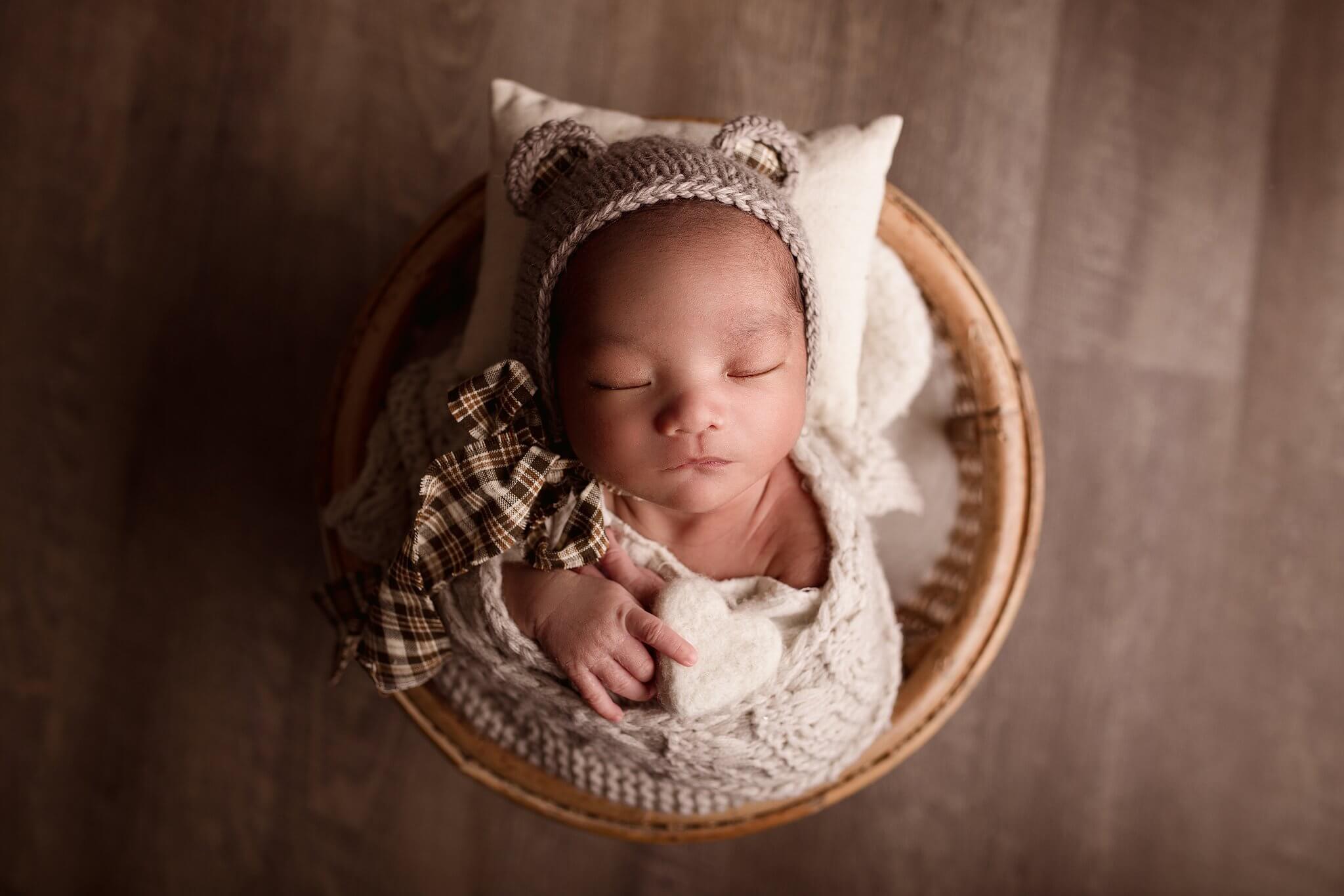 Adorable Newborn boy in a round basket wearing a teddy bear hat with plaid ties, holding a felt heart