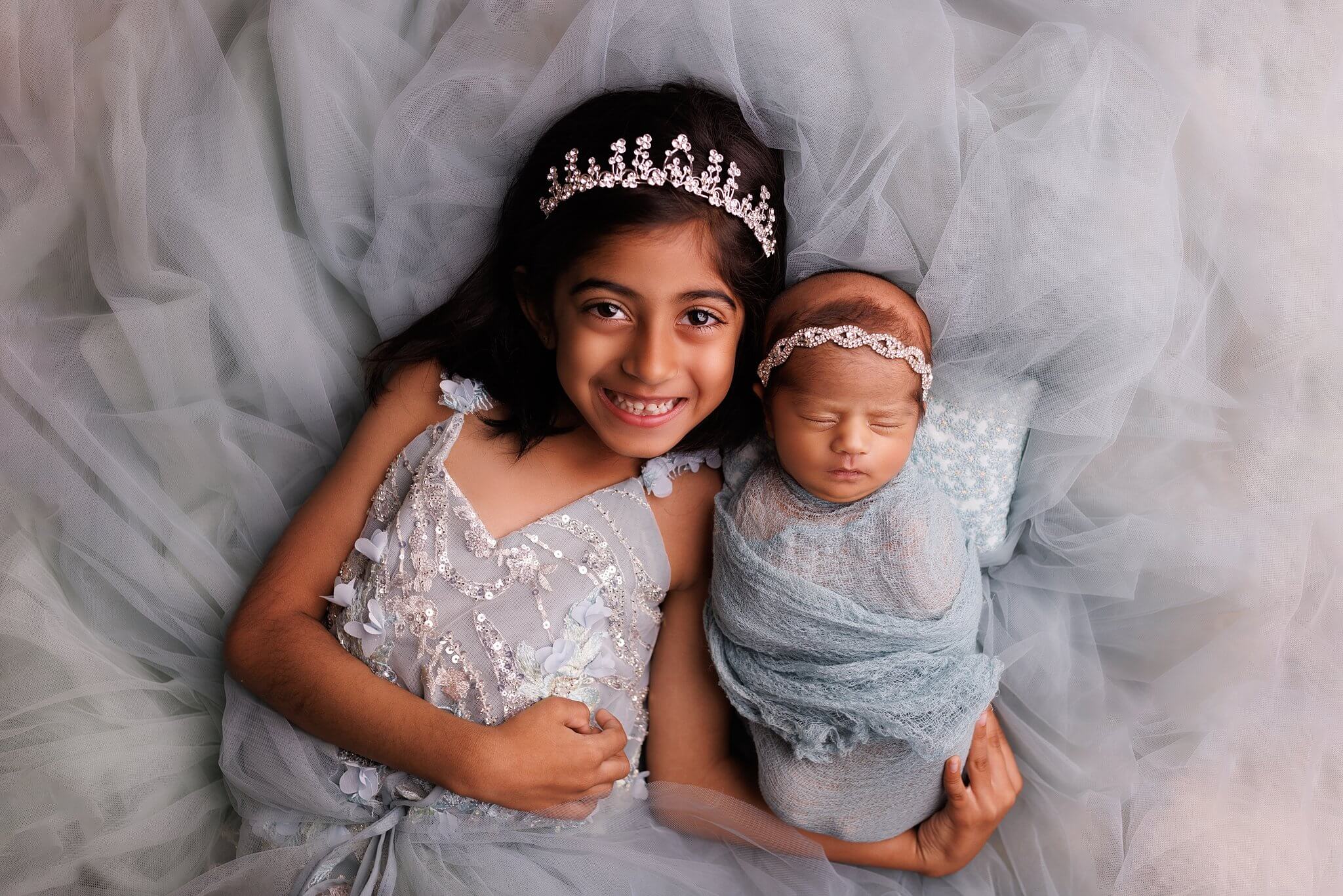 Sibling photo of a young girl in a blue gown laying on her back holding her newborn baby sister