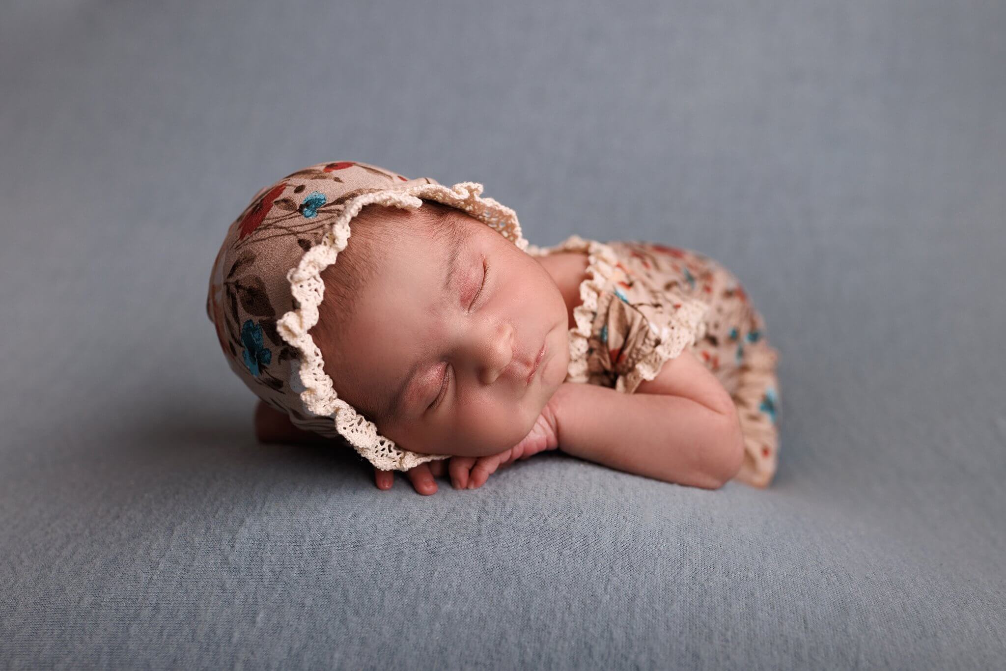 Newborn baby girl on a blue backdrop in a floral print bonnet and outfit by Toronto Newborn Photographer