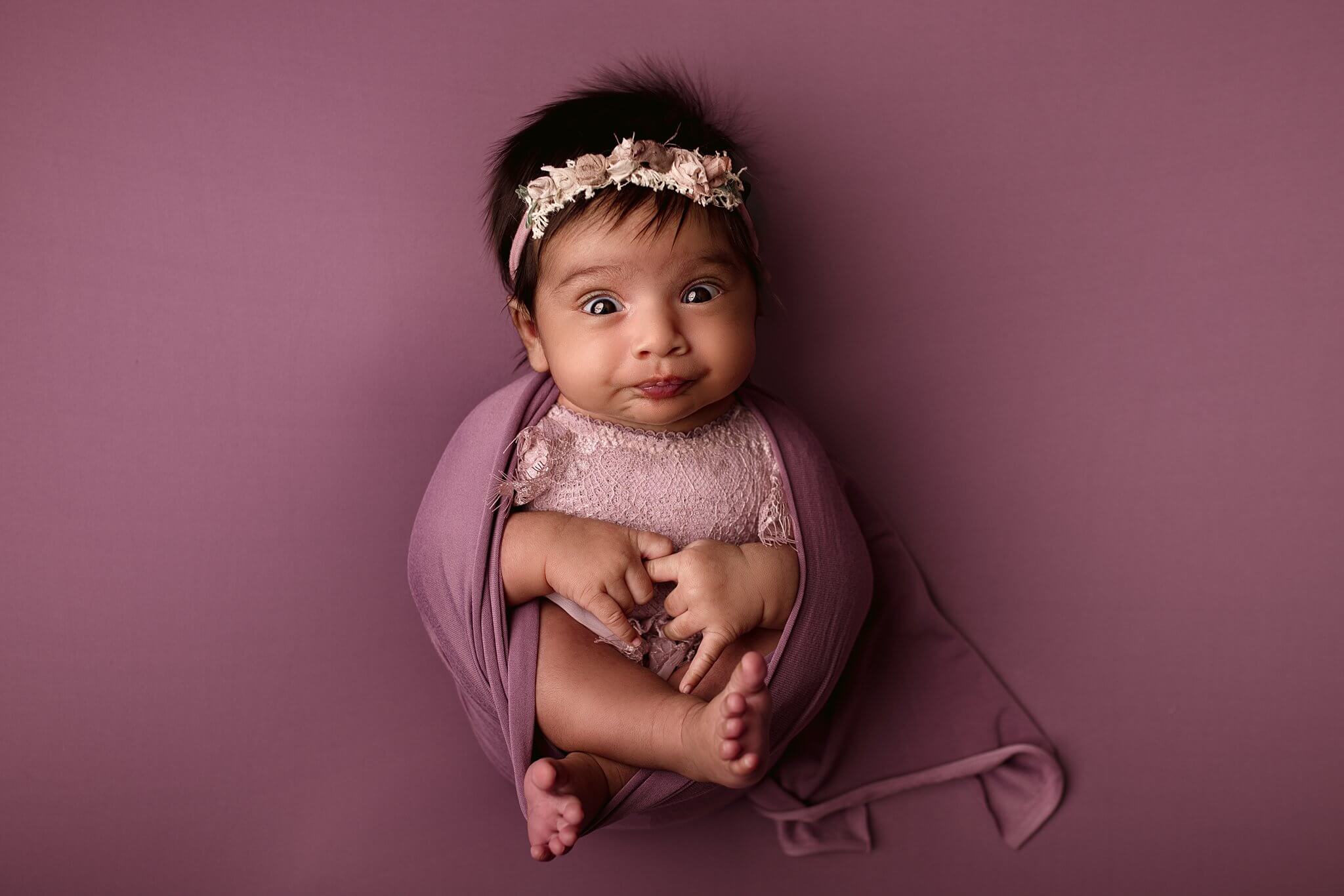 Awake and surprised looking newborn girl on lilac fabric with a floral headband