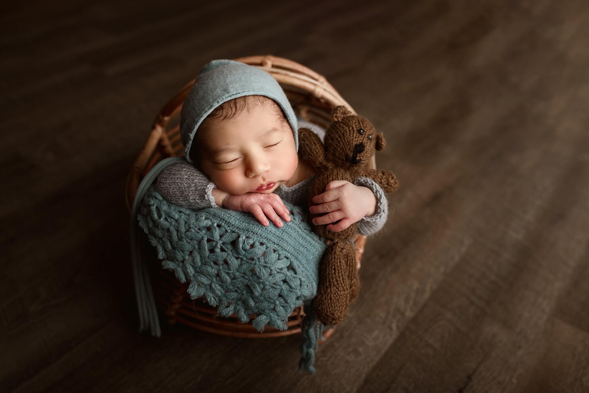 Newborn baby boy in a basket and holding a teddy bear in ocean colored bonnet
