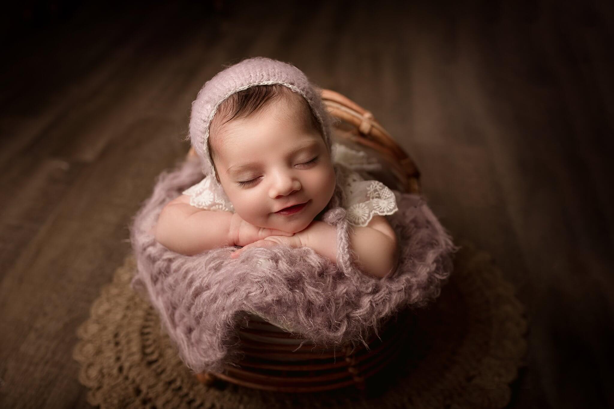 Newborn Baby Girl smiling asleep in a basket on lavender blanket with matching lavender bonnet by Toronto Newborn Photographer