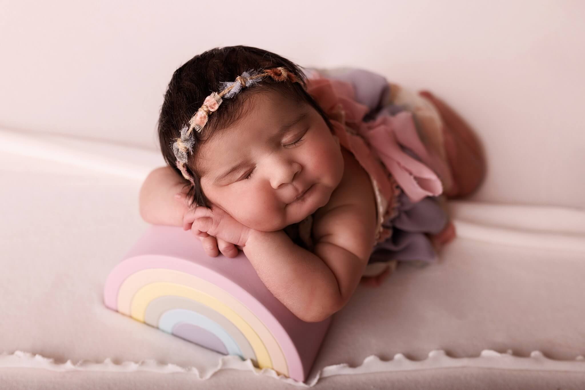 Newborn baby girl wearing a small bow rainbow headband, resting on a rainbow prop