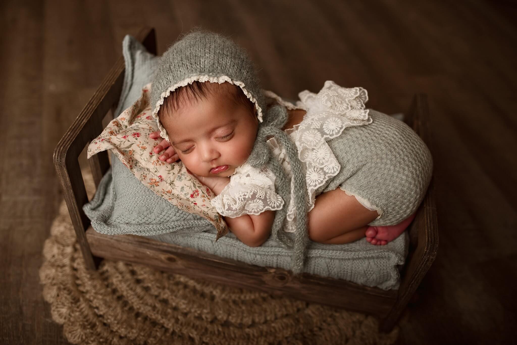 Newborn girl in a lace and sage outfit and matching sage bonnet on a tiny bed