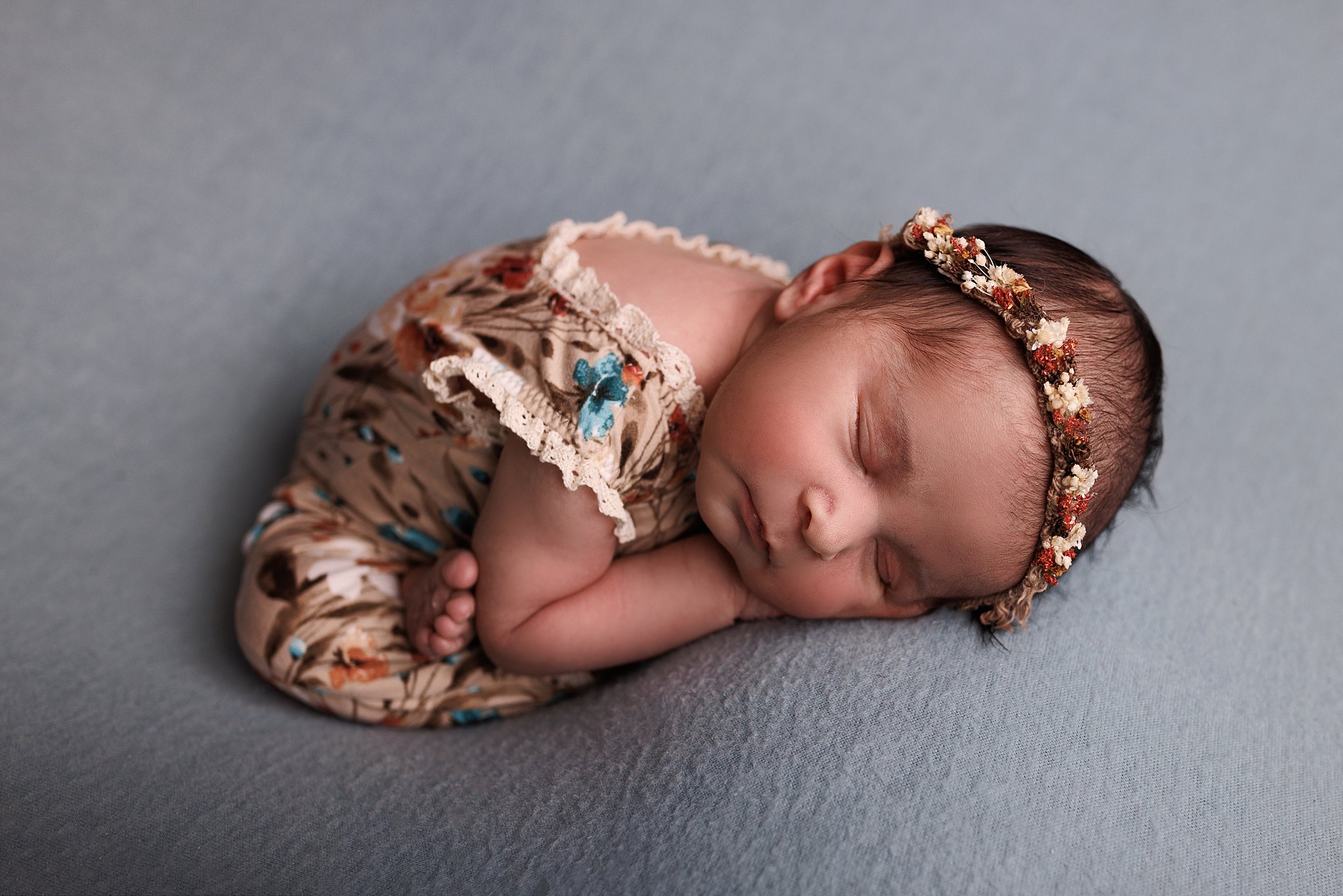 A newborn wearing a floral onesie with lace edges sleeps with legs crossed in a studio Toronto baby clothes
