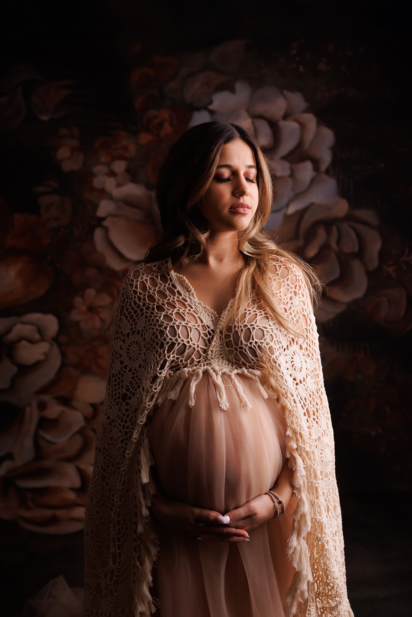 mother-to-be stands in a woven shawl and maternity gown in a studio with a flower backdrop Toronto prenatal massage
