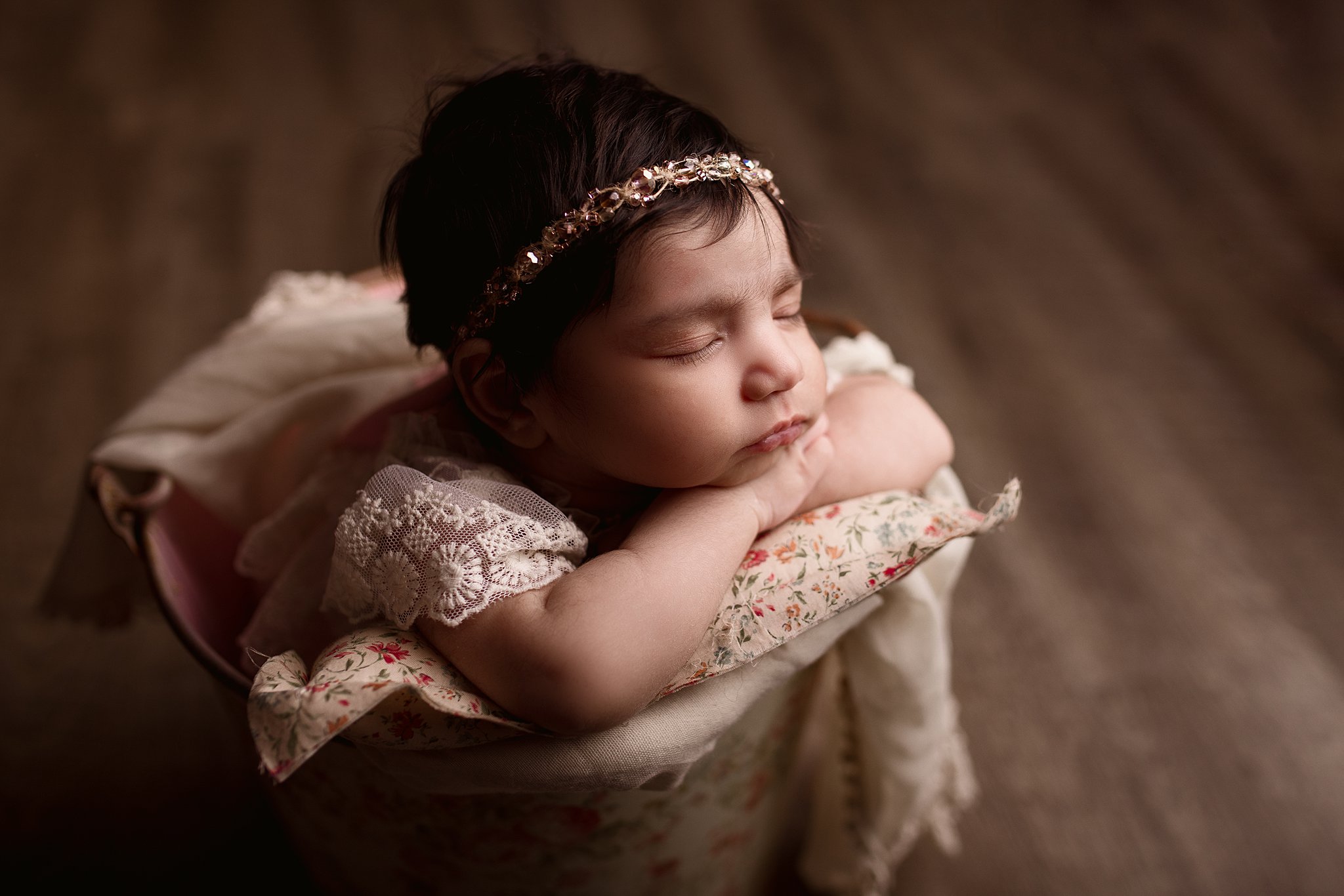 A newborn baby sleeps resting her head on her hands in a white lace dress inside of a tin bucket