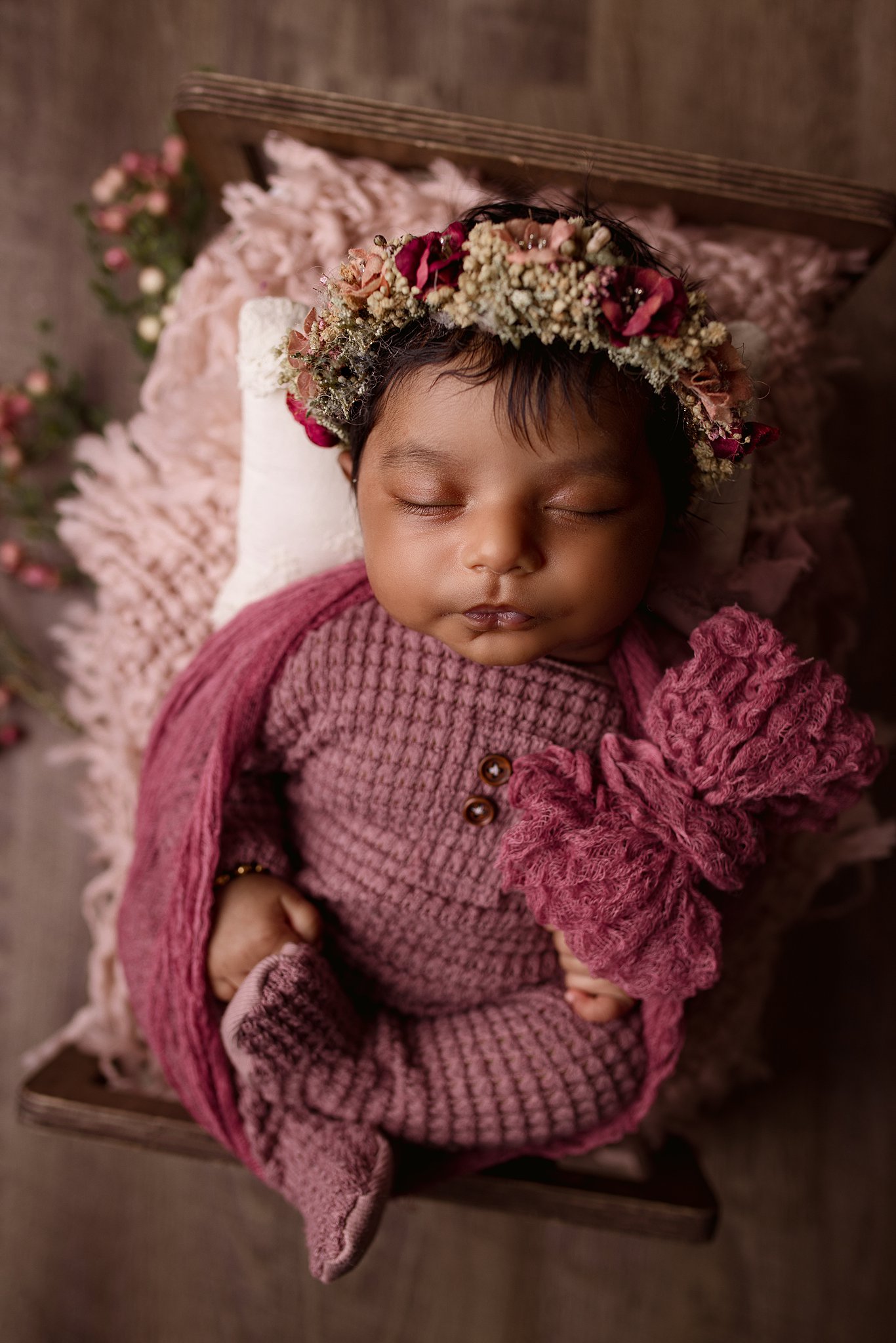A newborn baby sleeping in a knit purple onesie in a tiny wooden bed with a flower headband Ava's Appletree