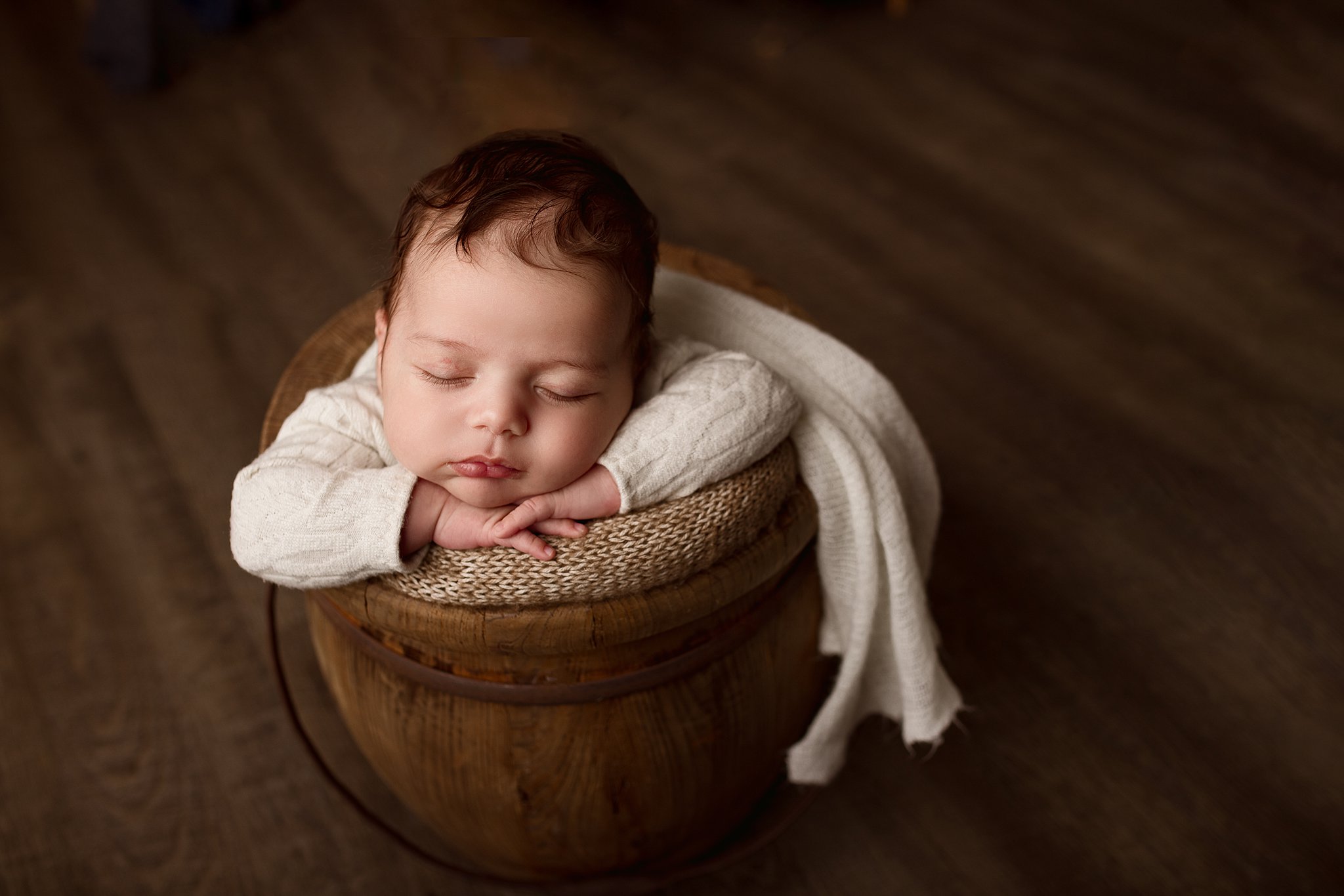 A newborn baby sleeps in a hardwood bucket wearing a white sweater resting head on it's hands
