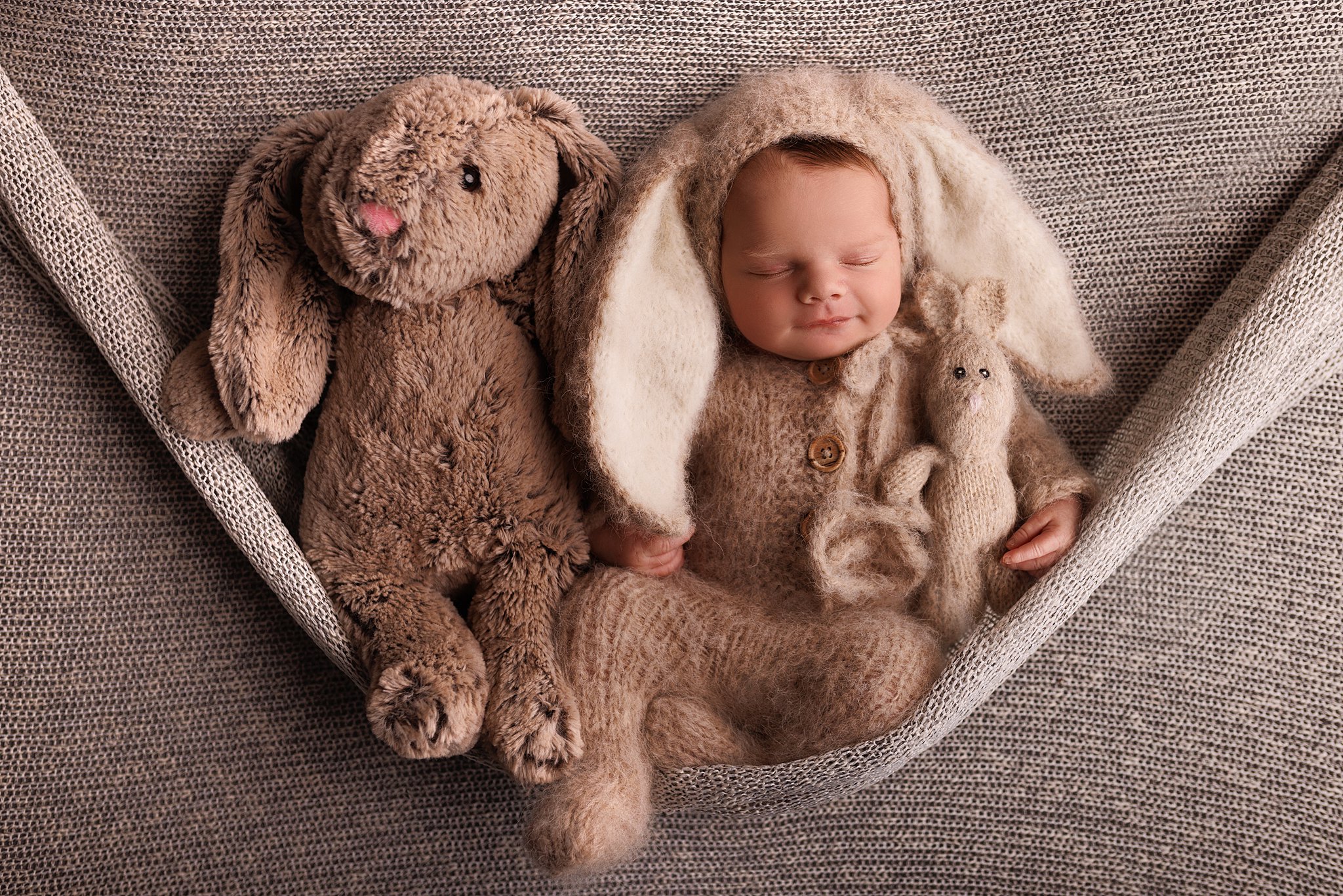 A newborn baby sitting on a knit sling sleeps wearing a brown knit bunny suit with a two stuffed bunnies of the same color baby furniture vaughan