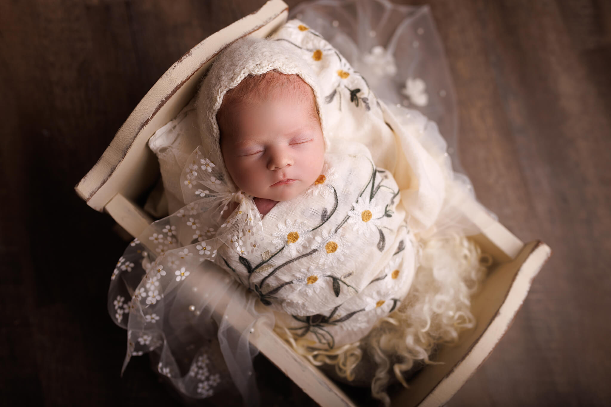 A newborn baby sleeps wrapped in a daisy blanket in a wooden crib and white bonnet