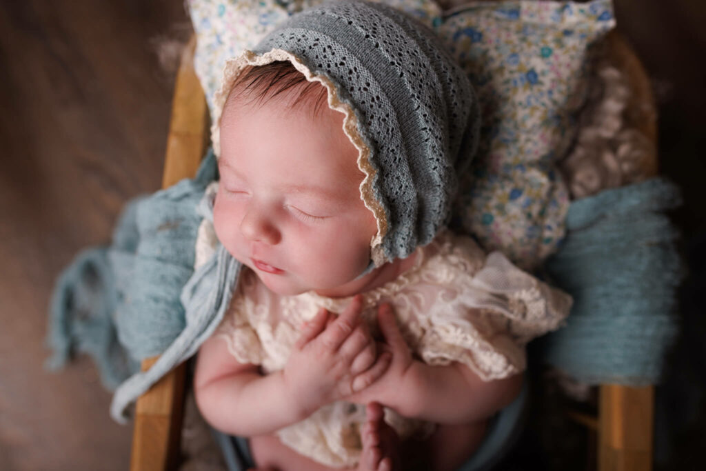 A newborn baby in a lace dress and blue bonnet sleeps in a wooden bed blanchou