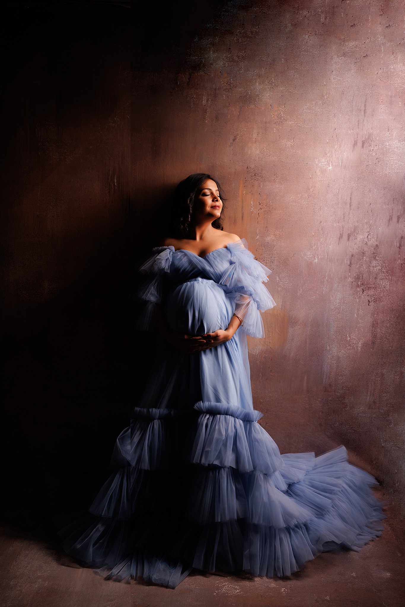 A mother to be in a blue maternity gown leans against a wall in a studio