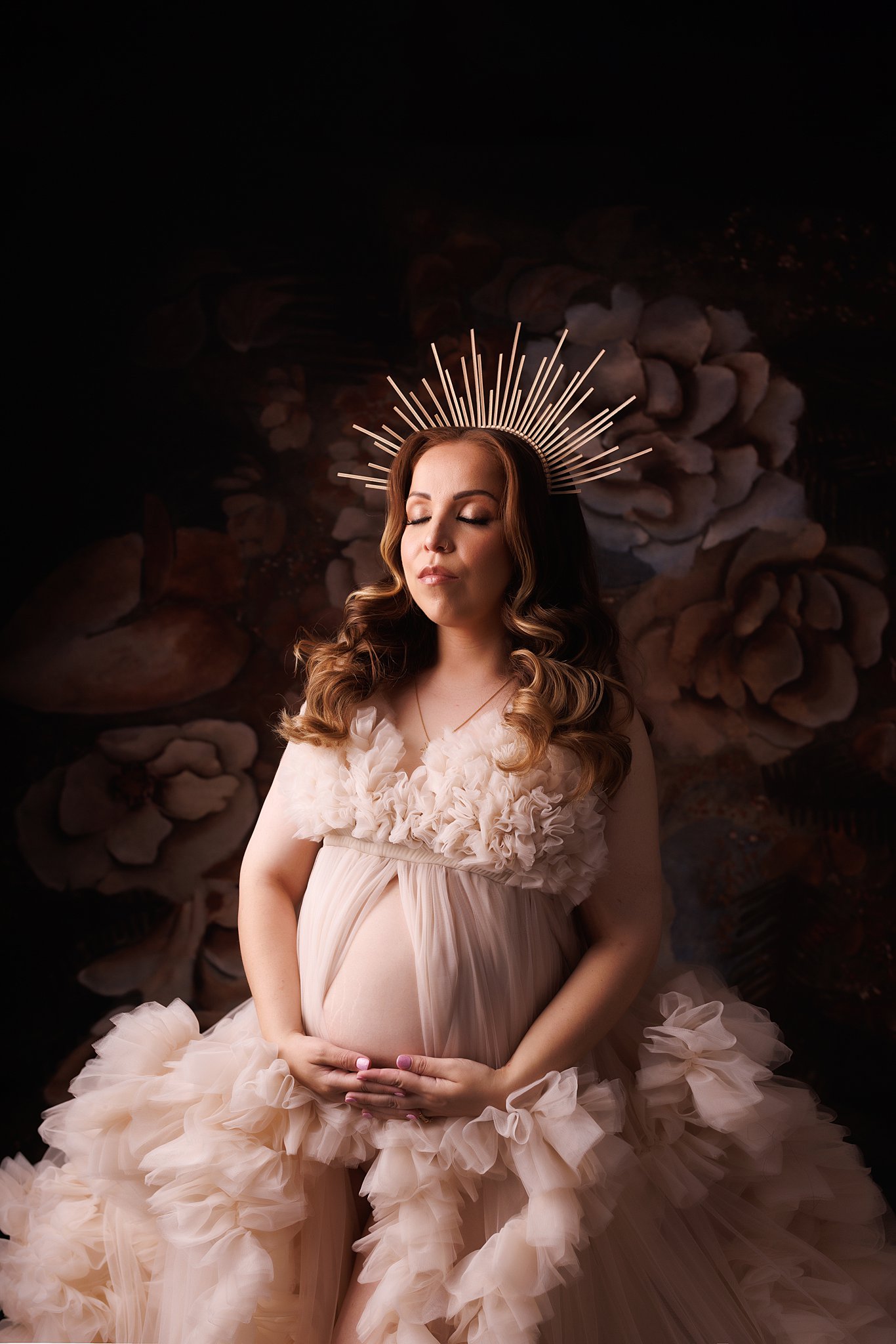 A mother to be in a pink maternity gown sits in a studio in a sun crown