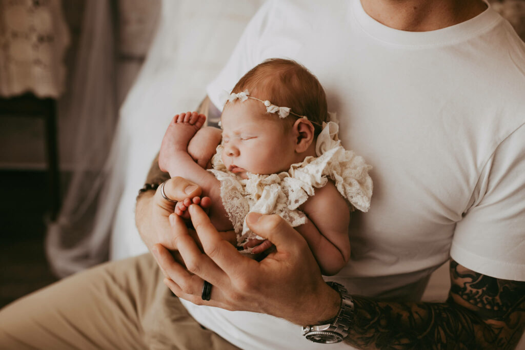 A father holds his sleeping newborn baby curled up against his chest in a white dress