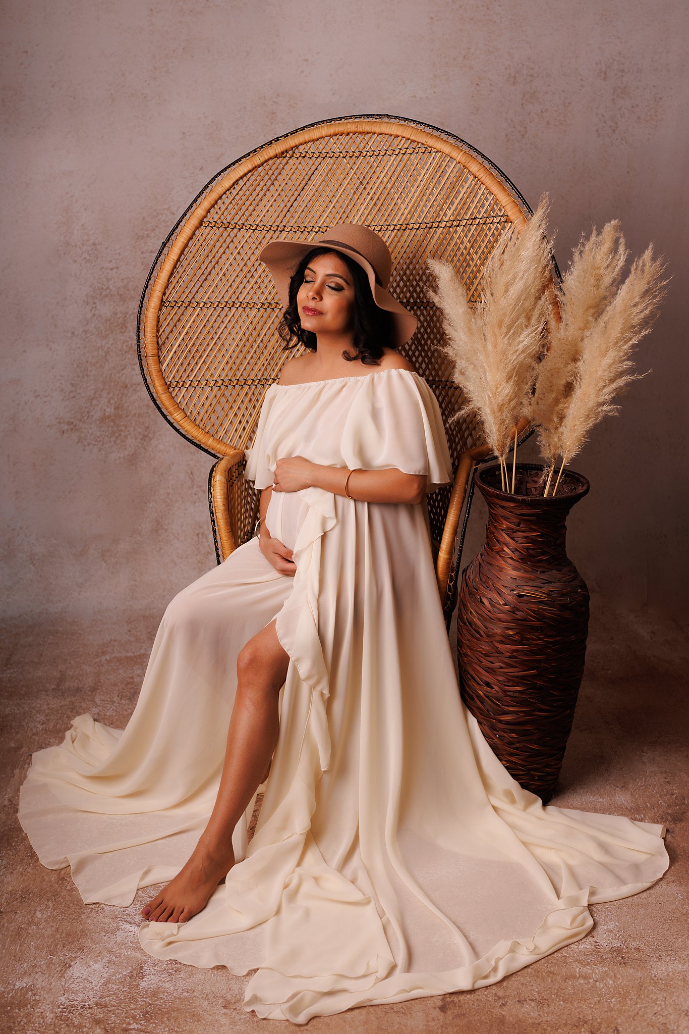 A mother to be in a long white maternity gown sits in a tall wicker chair in a studio cherry blossom doulas