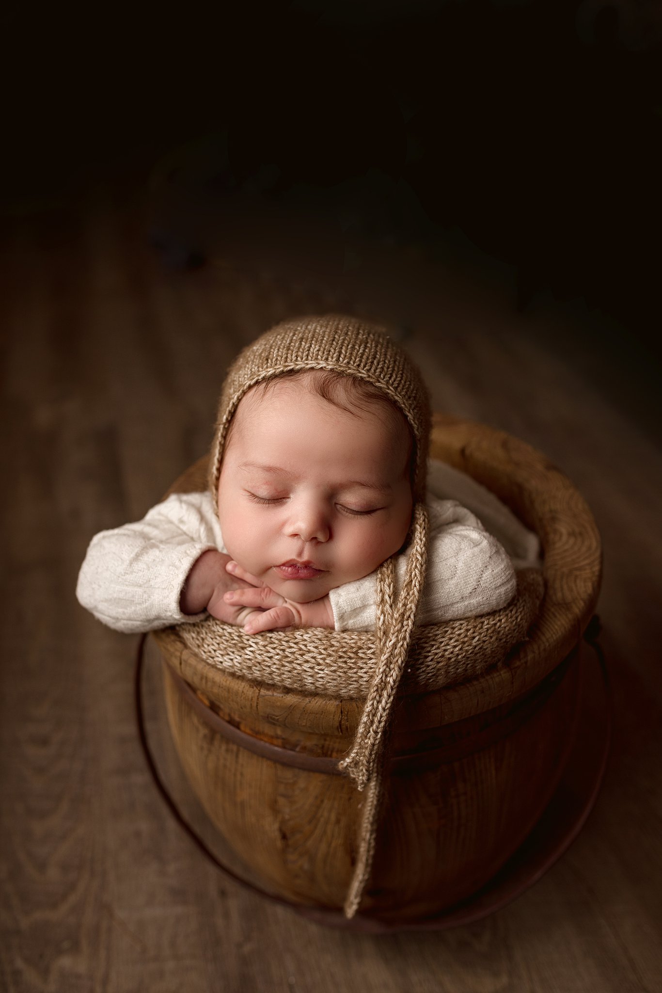 A newborn baby sleeps in a wooden bucket with head on its hands in a white sweater and brown knit hat diaper service toronto
