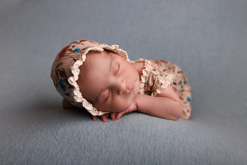 A newborn baby sleeps on a grey bed wearring a floral onesie with matching bonnet diaper service toronto