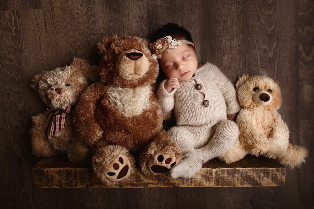 A newborn baby in a knit onesie sleeps on a wooden shelf with three teddy bears
