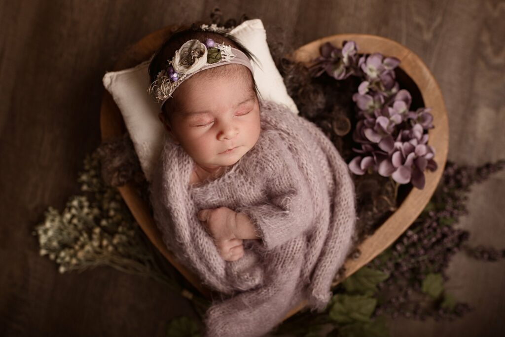 A newborn baby sleeps while wrapped in a purple knit blanket in a basket of purple flowers macklem's toronto