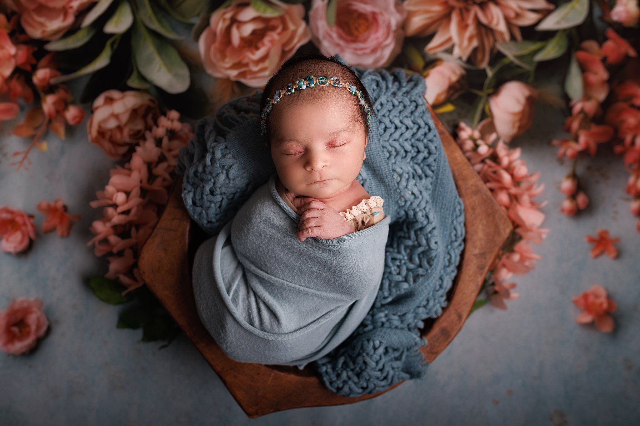 A newborn baby sleeps in a blue swaddle with a matching blanket in a wooden bucket