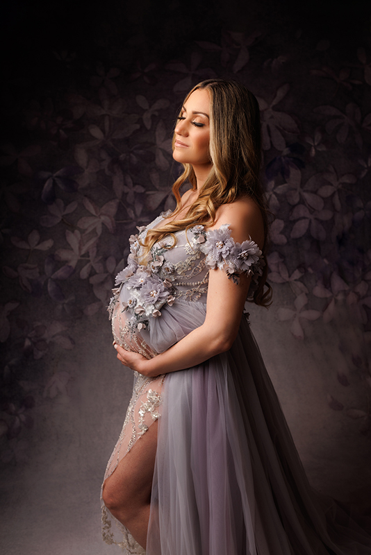 Beautiful Mother to be in a lavender gown with flowers in front of a coordinating lavender backdrop